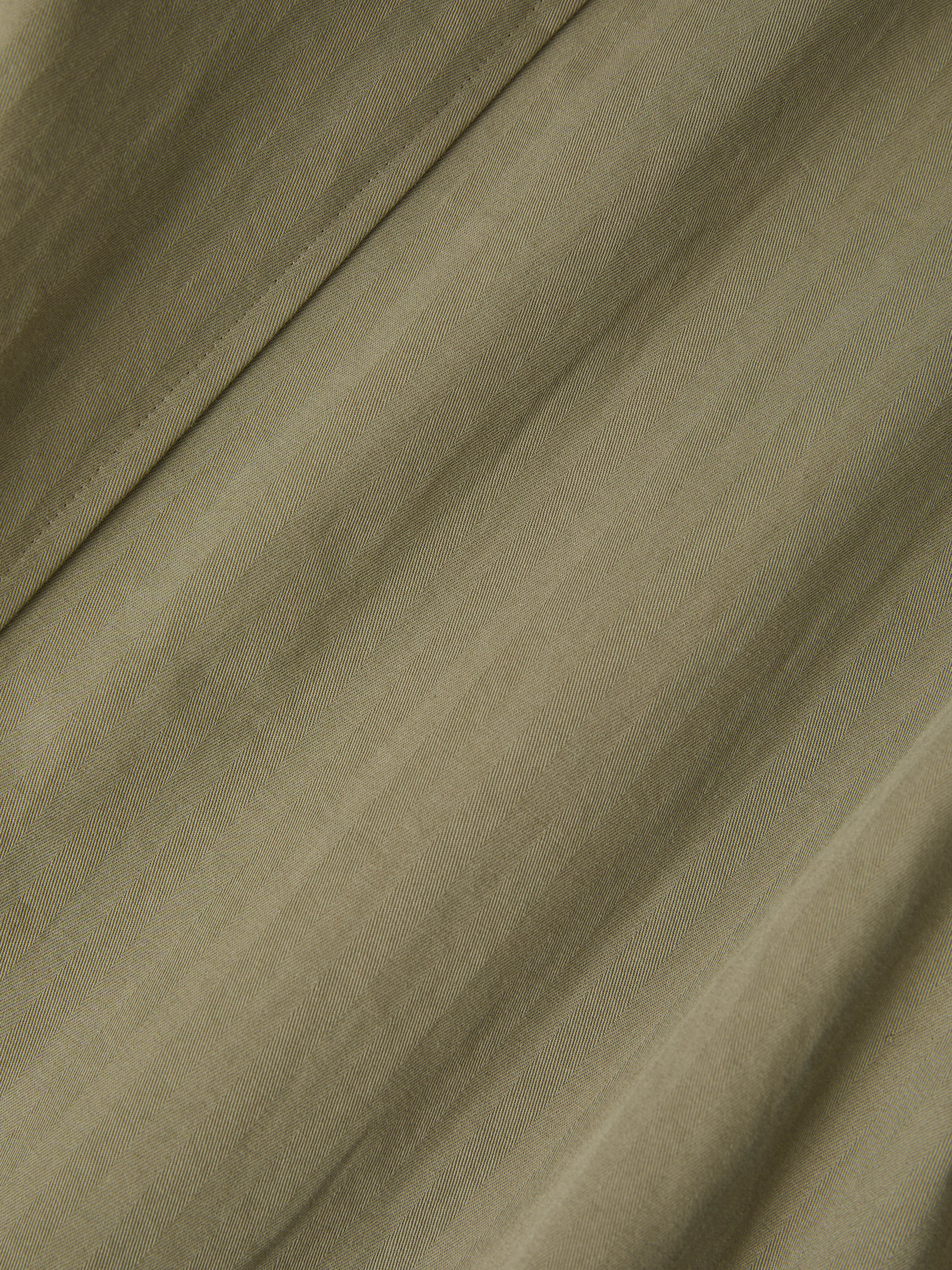 A close-up of smooth, olive green fabric with subtle diagonal folds mirrors the elegant texture of FOUND's Herringbone Twill Trench Coat.