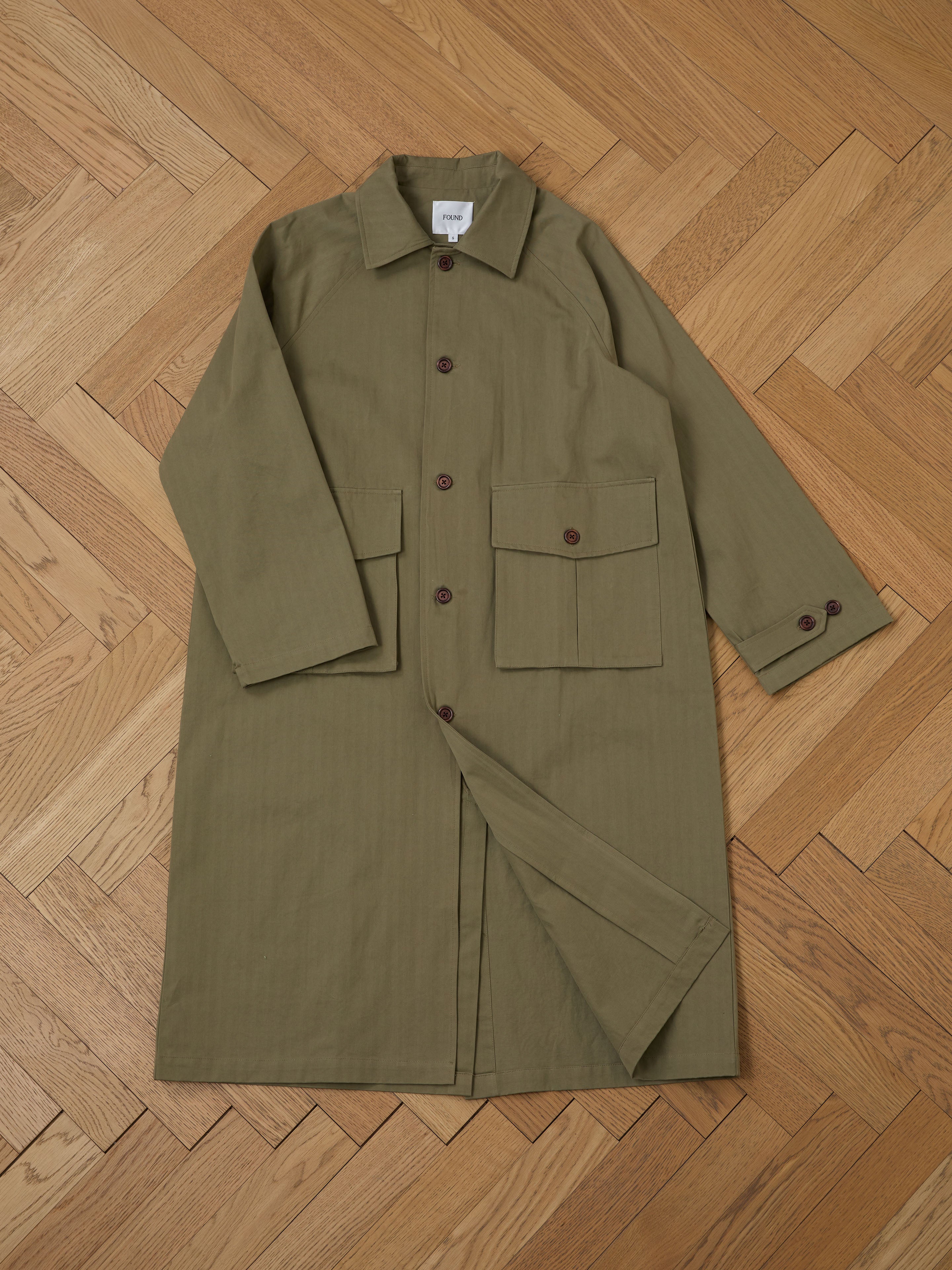 The Herringbone Twill Trench Coat by FOUND, featuring a relaxed fit in olive green with brown buttons and utility pockets, is elegantly displayed on a wooden herringbone floor.