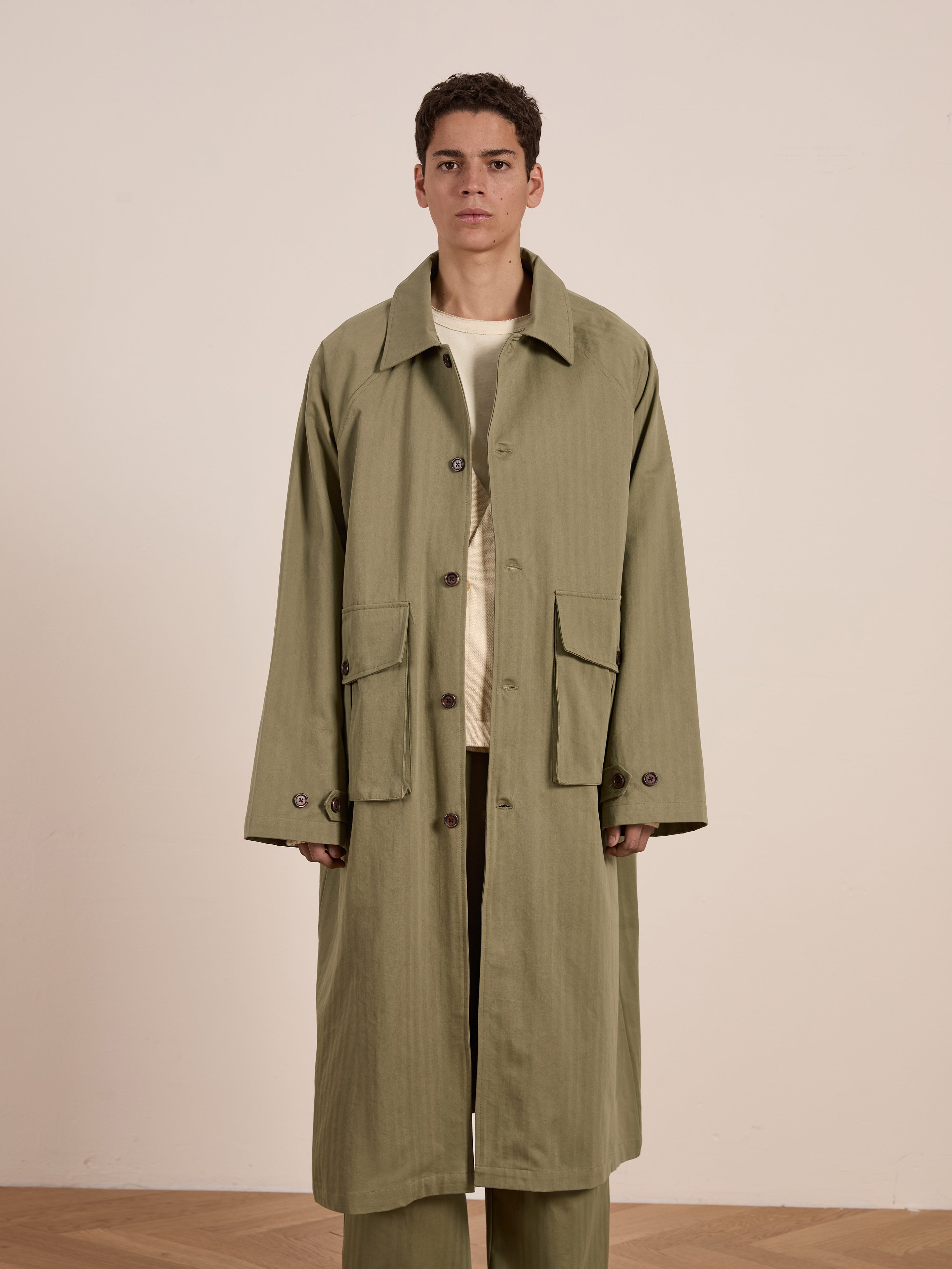 A person models the FOUND Herringbone Twill Trench Coat, featuring a relaxed fit, utility pockets, and large buttons, in a long green design against a plain background.