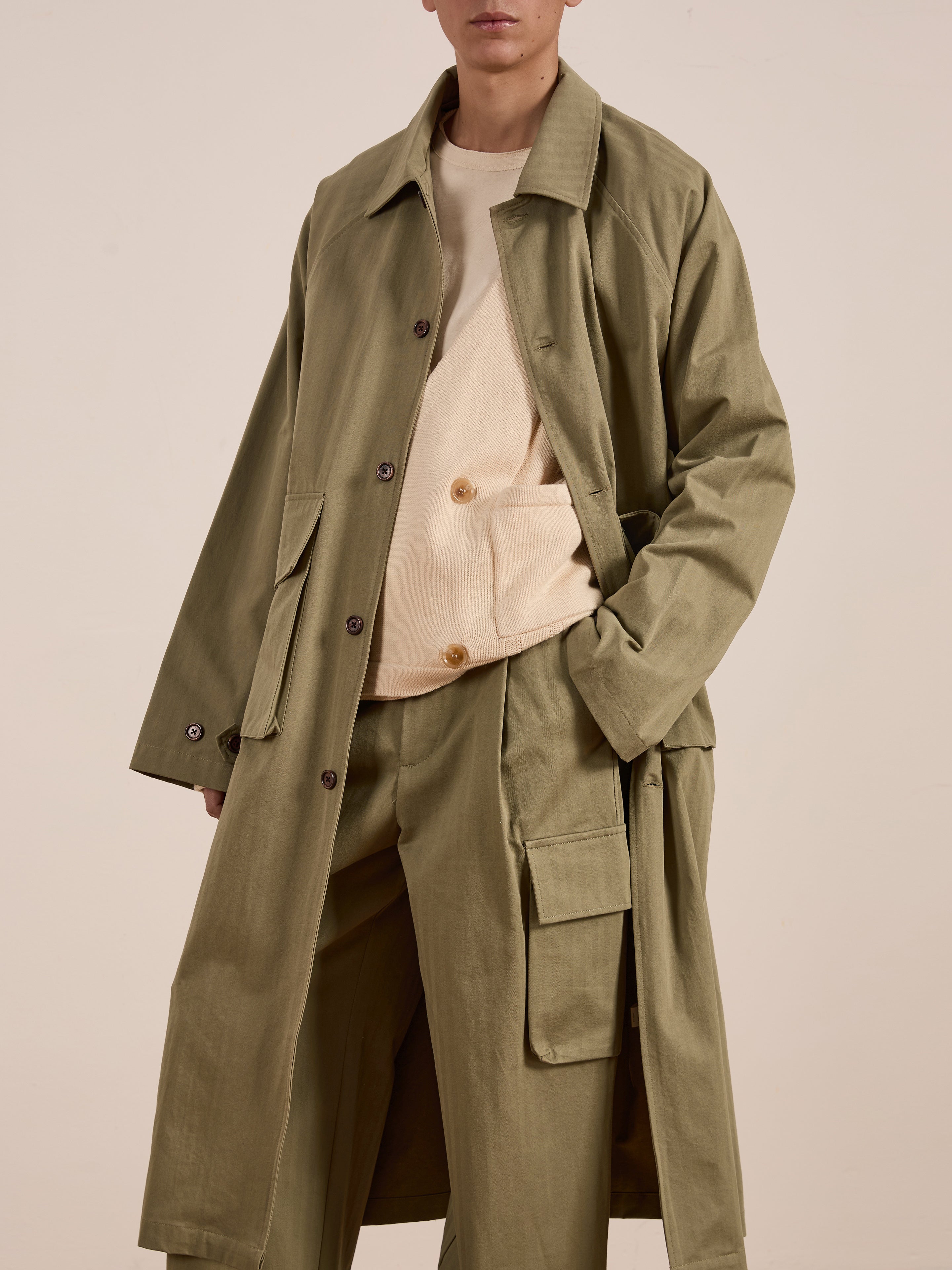 A person stands against a plain background in FOUND's olive green Herringbone Twill Trench Coat with matching pants and a beige top, highlighting a relaxed fit and utility pockets.