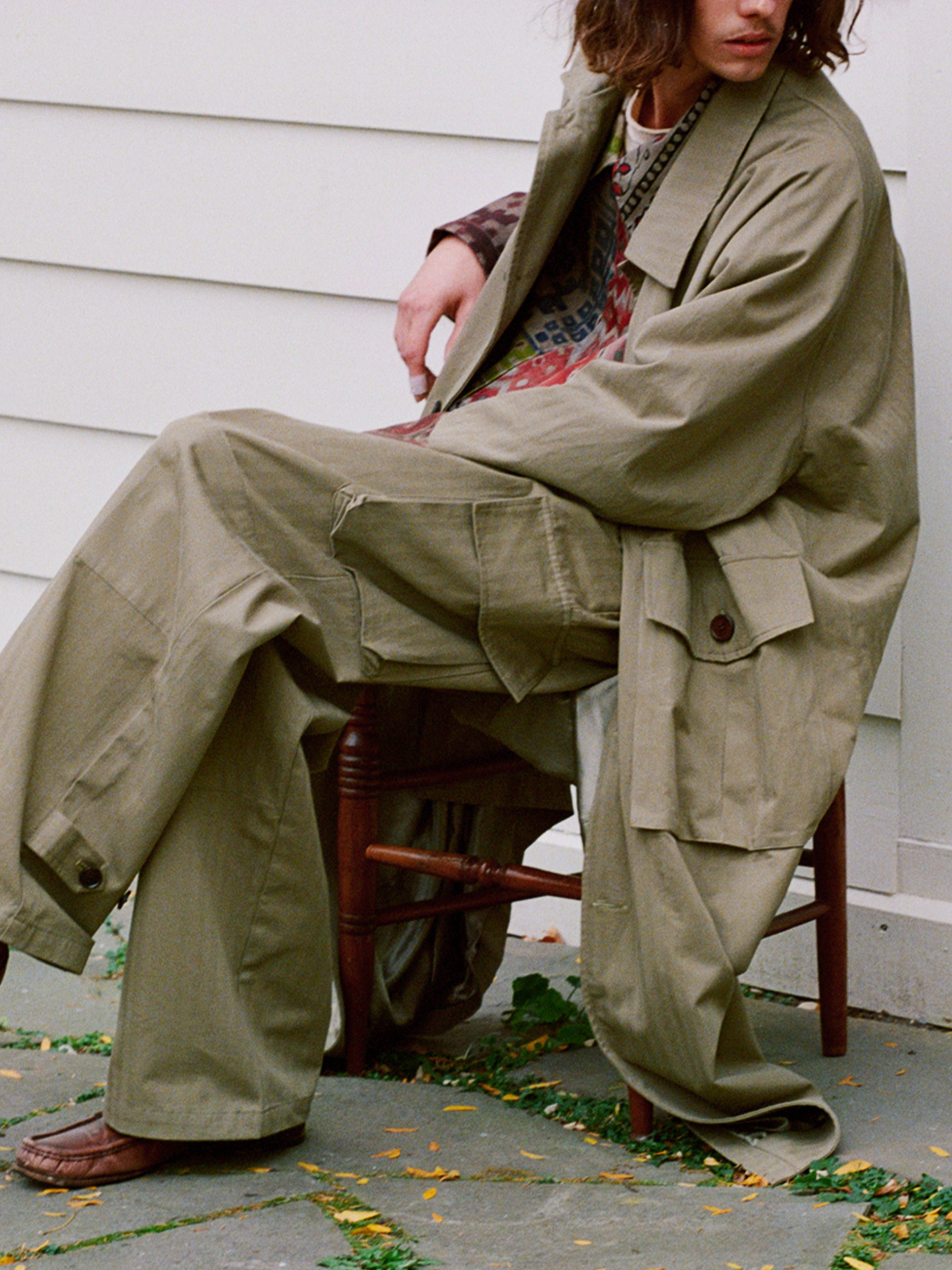 Seated outdoors on a chair, the person wears FOUND's Herringbone Twill Trench Coat with utility pockets, relaxed fit pants, and a colorful patterned inner shirt. Brown shoes peek below against a white wall background.