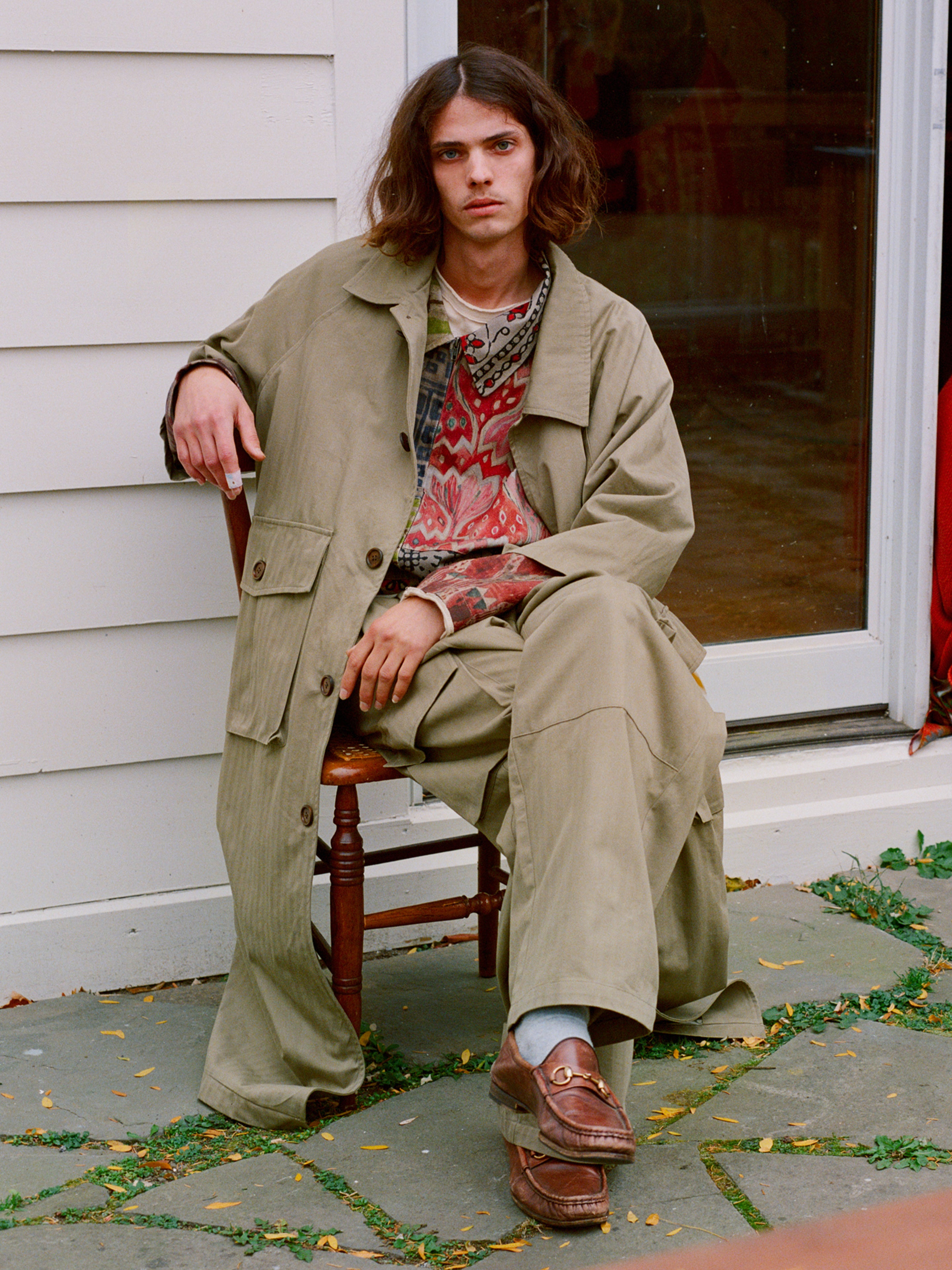 A person relaxes in a FOUND Herringbone Twill Trench Coat and patterned shirt, one leg crossed. The glass door and white wall backdrop subtly highlight the coat's utility pockets.