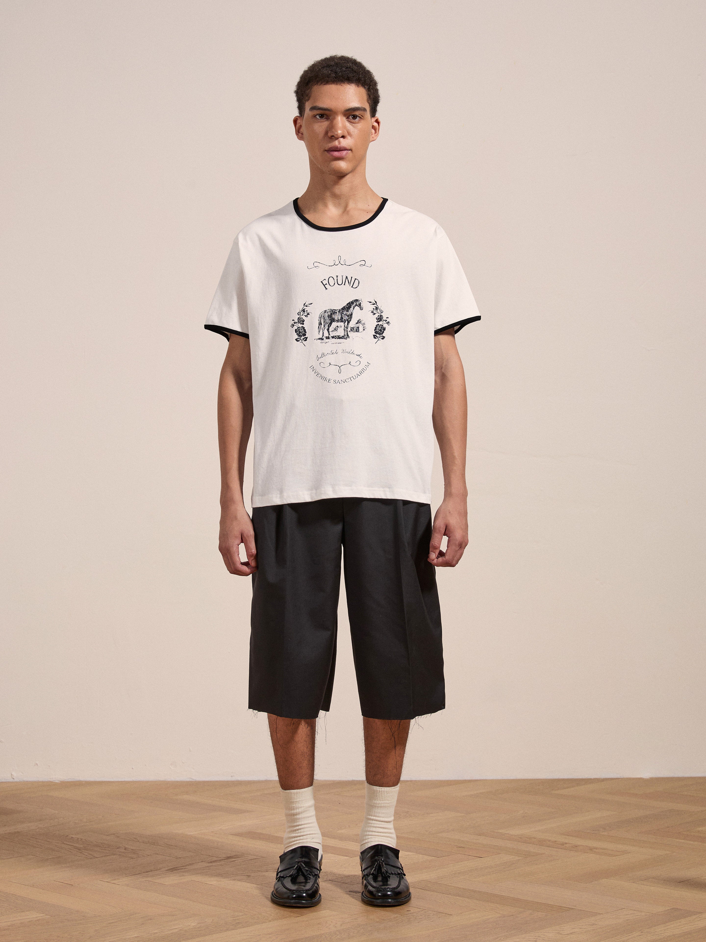 A person stands on a wooden floor wearing the Horse Crest Ringer Tee by FOUND, black knee-length shorts, white socks, and black shoes.