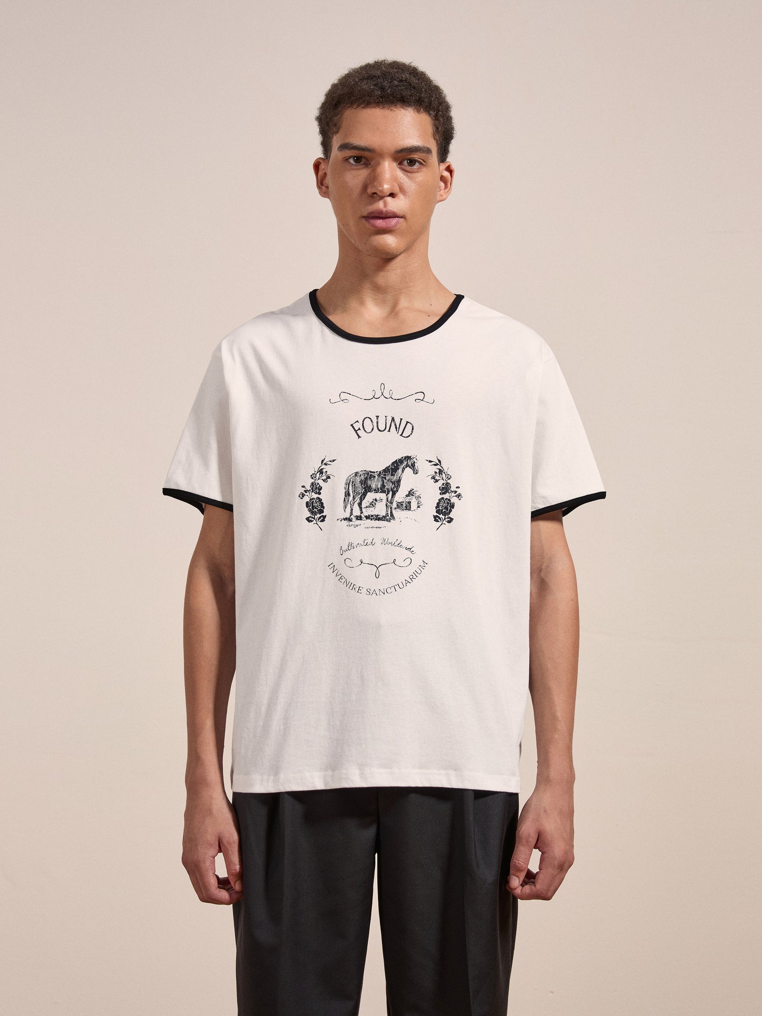 A young man wearing the FOUND Horse Crest Ringer Tee, featuring a white base with a black collar and an animal illustration on the front, stands against a plain background.