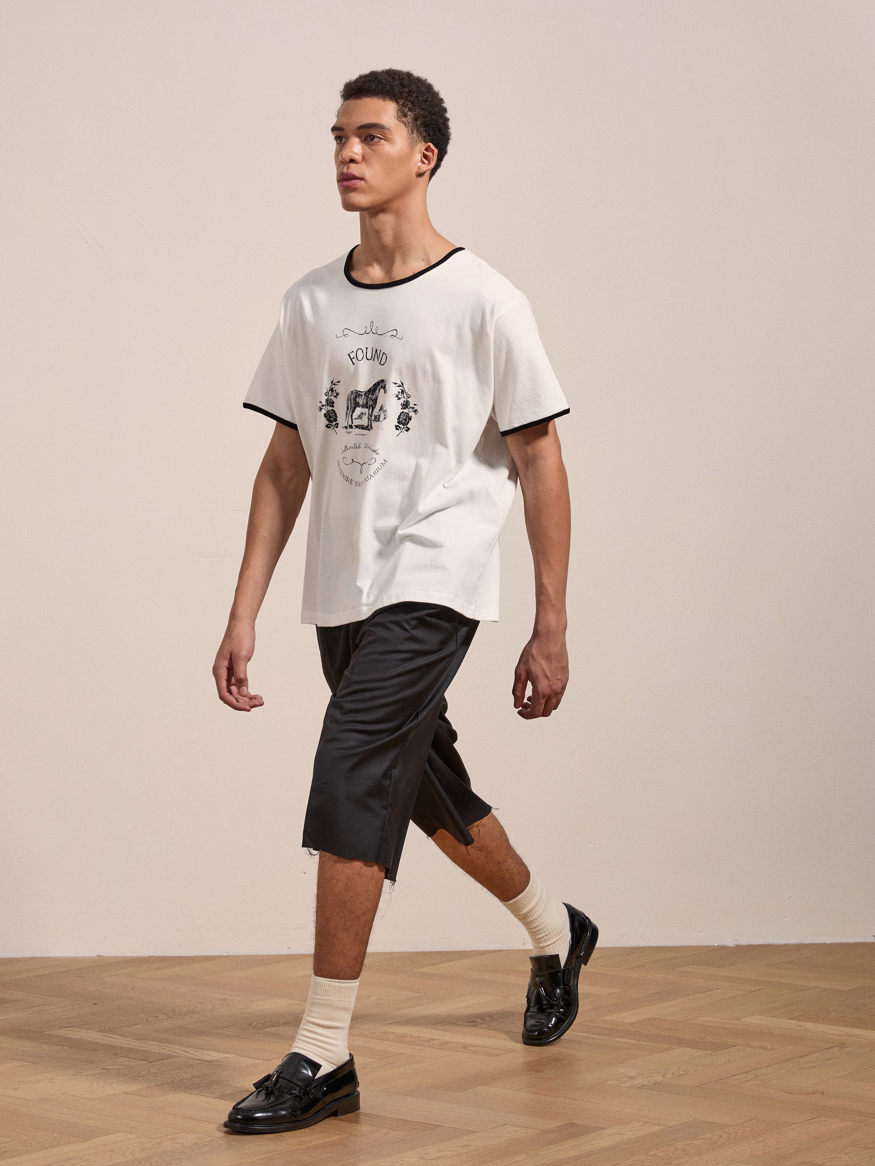 A man wearing the FOUND Horse Crest Ringer Tee, black knee-length shorts, white socks, and black dress shoes walks indoors with a neutral background.