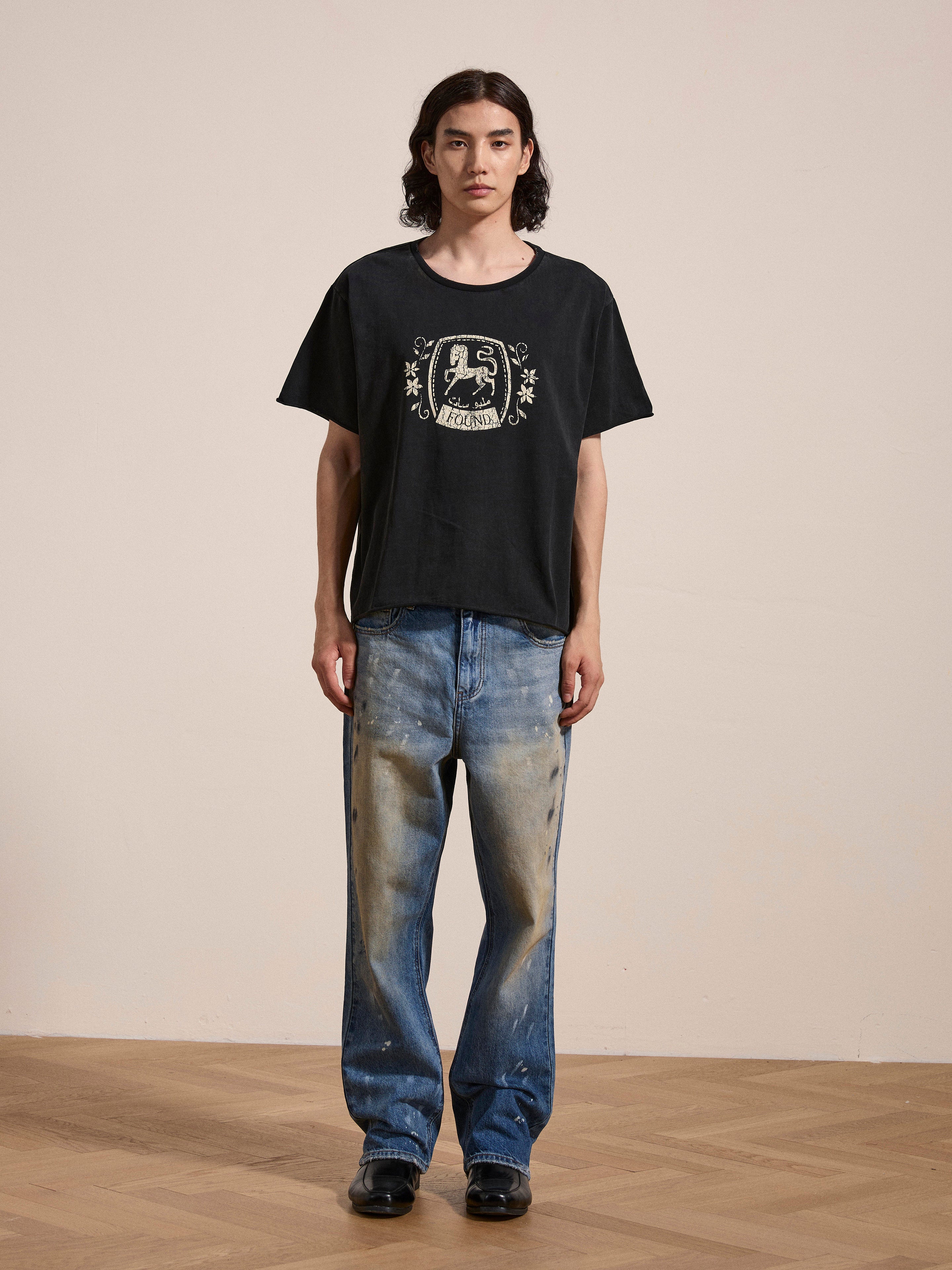 A person with long hair is wearing a black Horse Logo Print Tee from FOUND made of 100% cotton and distressed blue jeans, standing on a wooden floor against a plain beige wall.