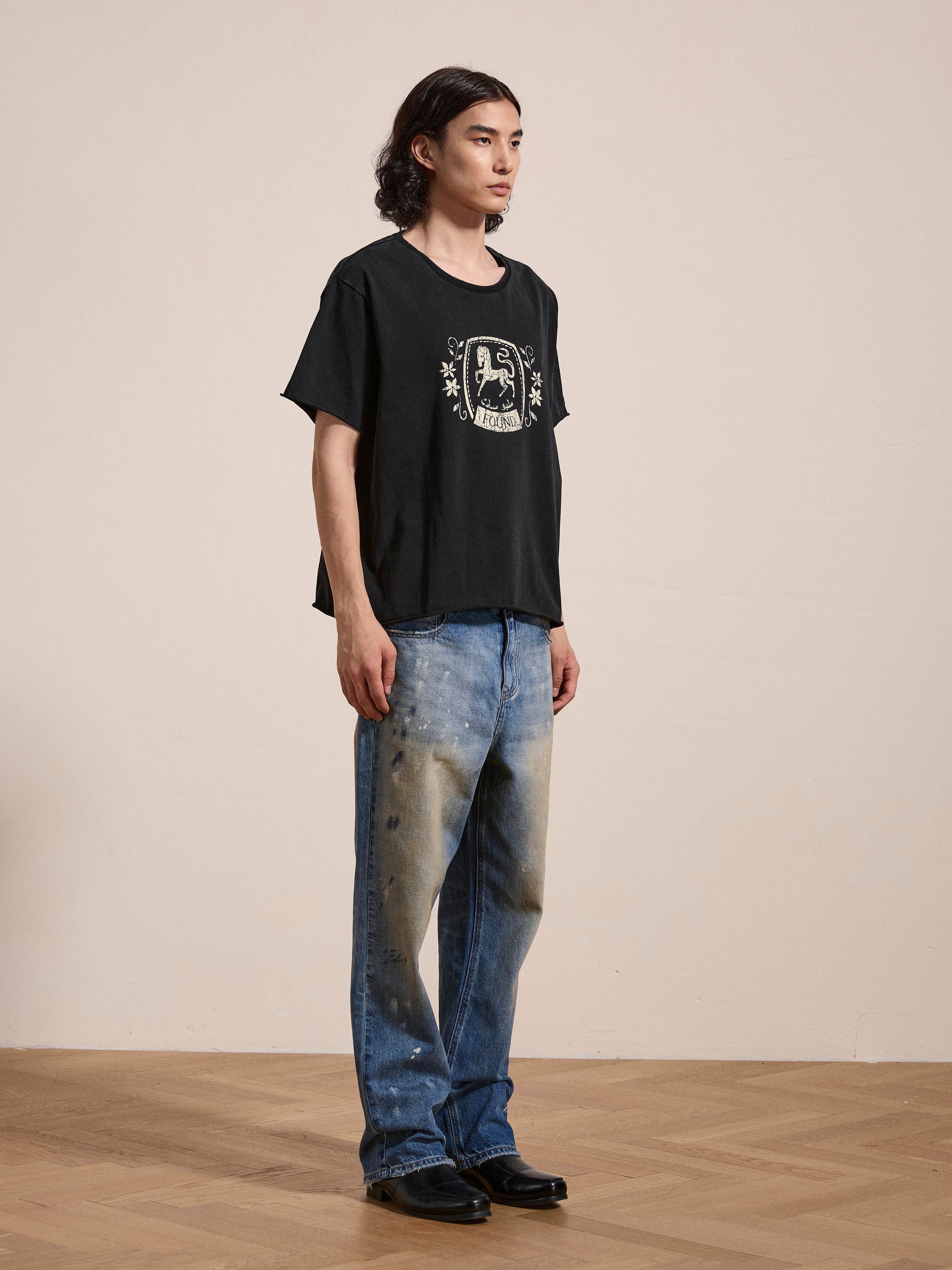 A person stands indoors wearing a FOUND Horse Logo Print Tee and distressed blue jeans with black shoes. The enzyme wash of the 100% cotton fabric gives the t-shirt a unique, worn look. The background is a plain beige wall and wooden floor.