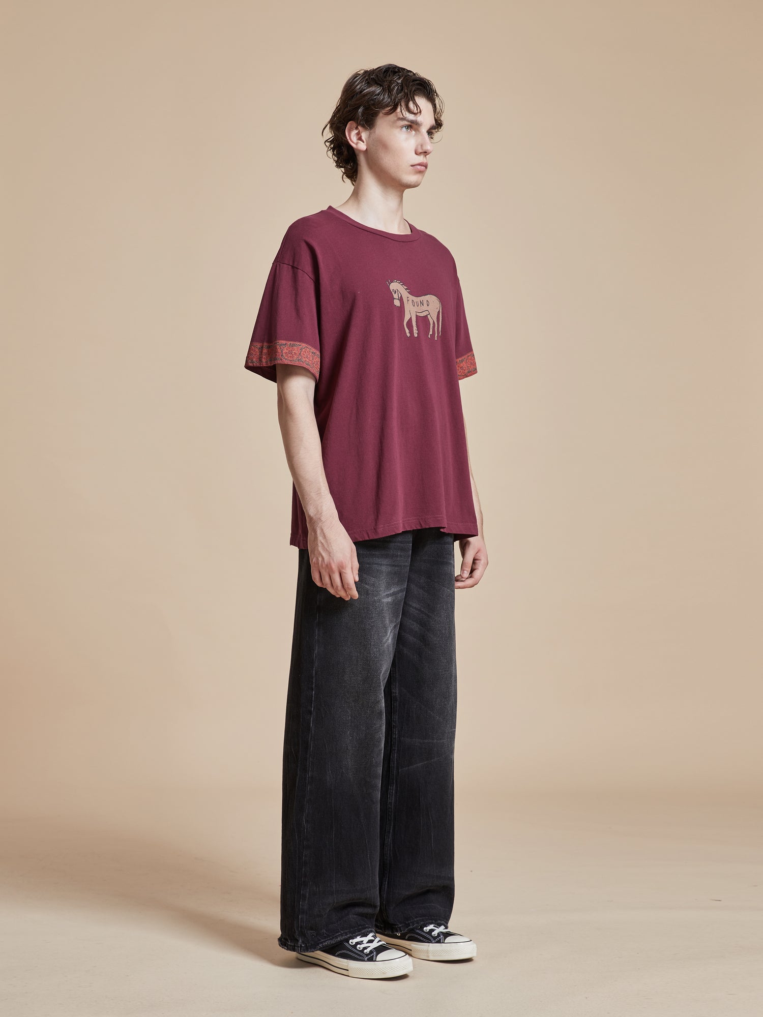 A man wearing a Found Horse Embellishment Tee burgundy t-shirt and black pants.