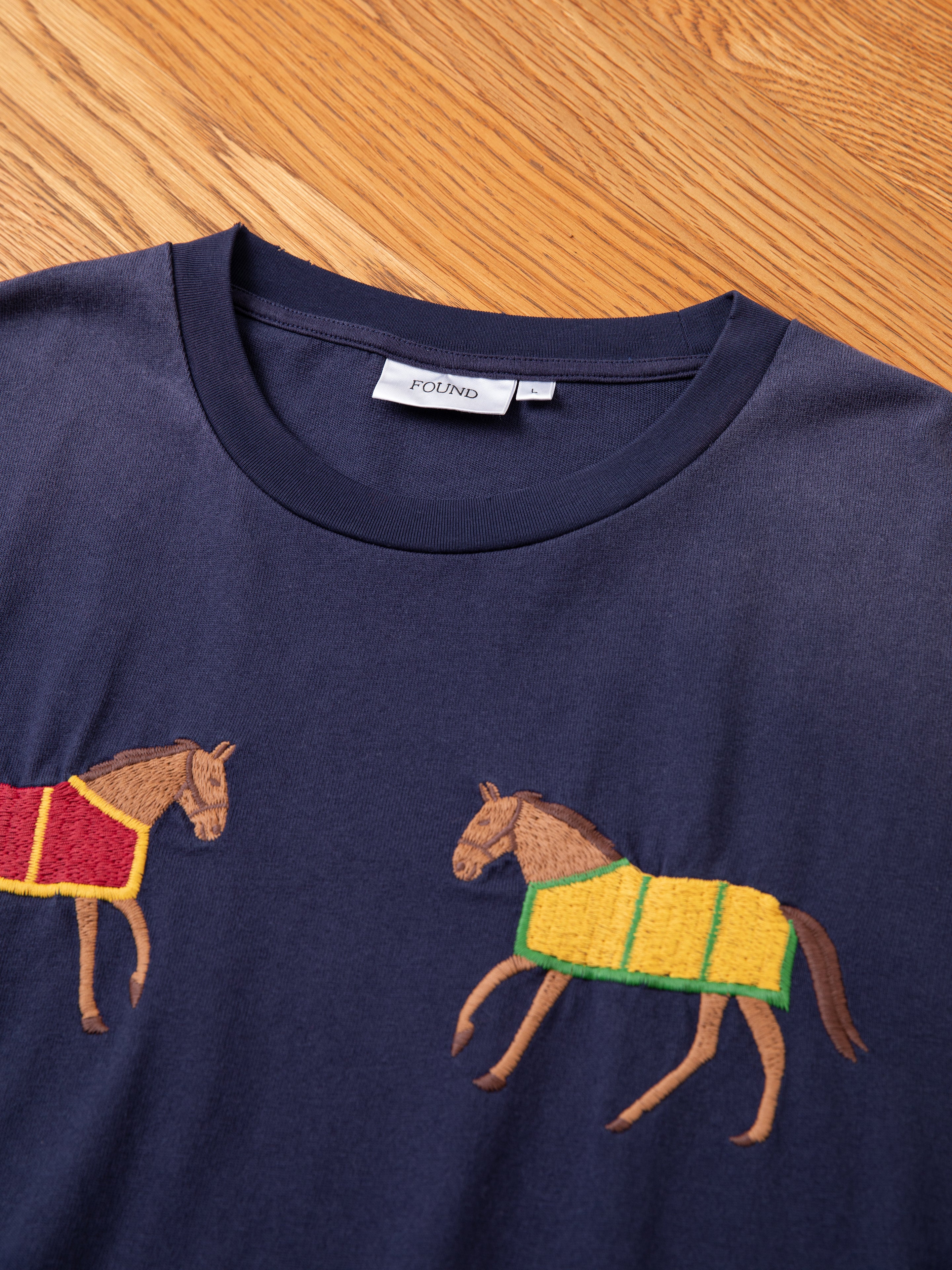 A Horse Equine Tee by FOUND, featuring embroidered horses—one with a red and yellow blanket and the other with a green and yellow blanket—rests on a wooden surface.