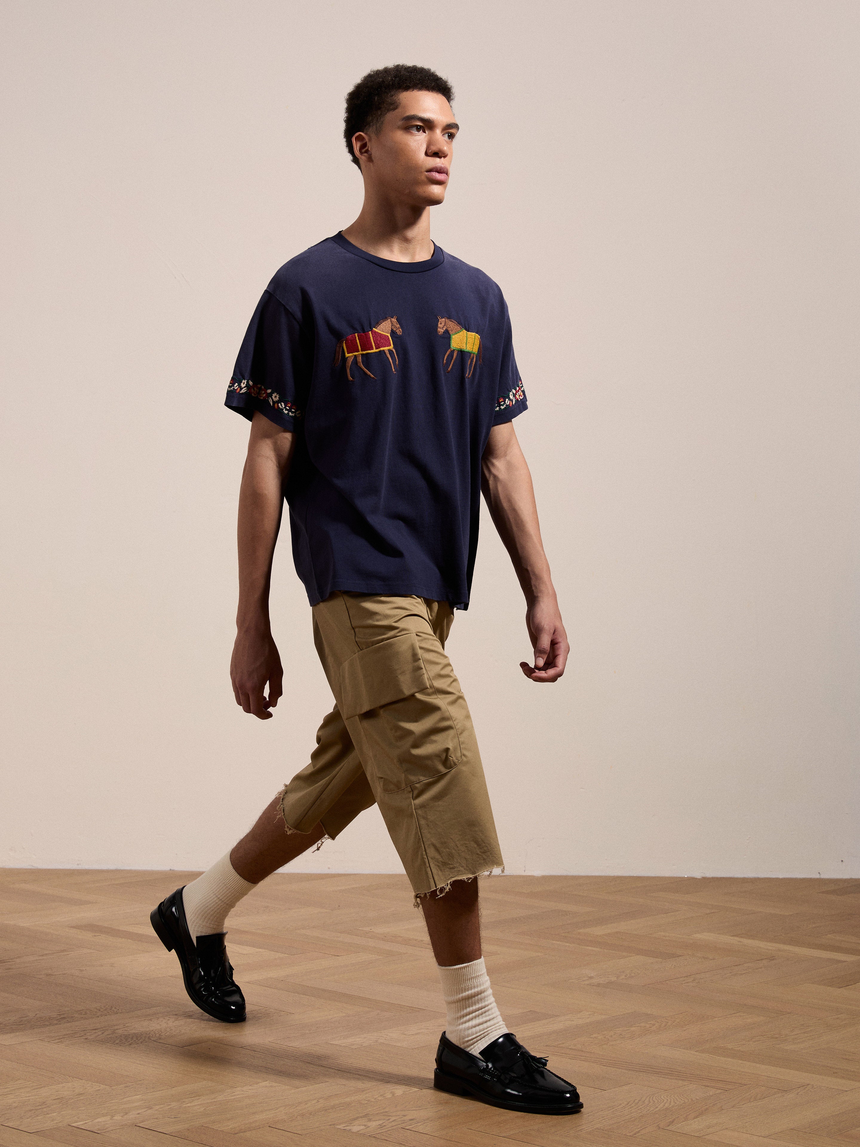 A person is walking indoors wearing a FOUND Horse Equine Tee, beige cargo shorts, white socks, and black shoes. The background features light-colored walls and a wooden floor.