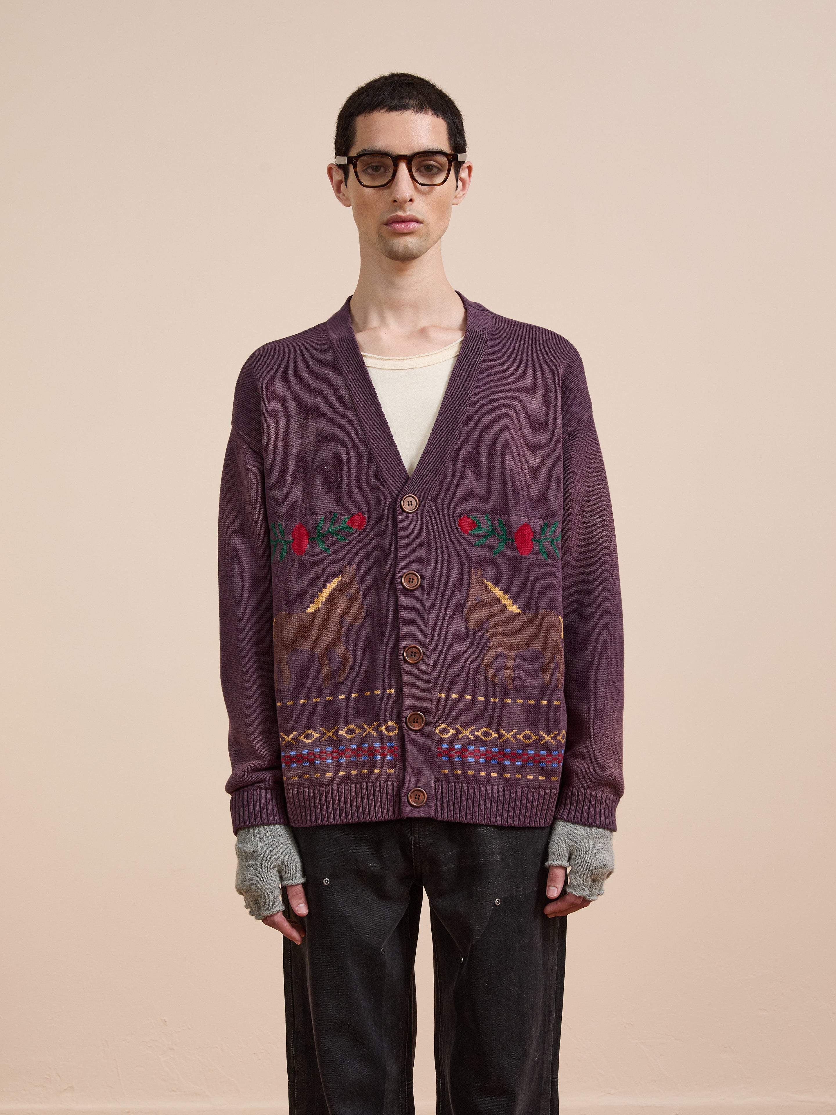 A person wearing glasses stands against a plain background in Found’s Horse Jacquard Cardigan, a purple unisex piece with animal and horse motifs, styled with a cream shirt, fingerless gloves, and dark pants.