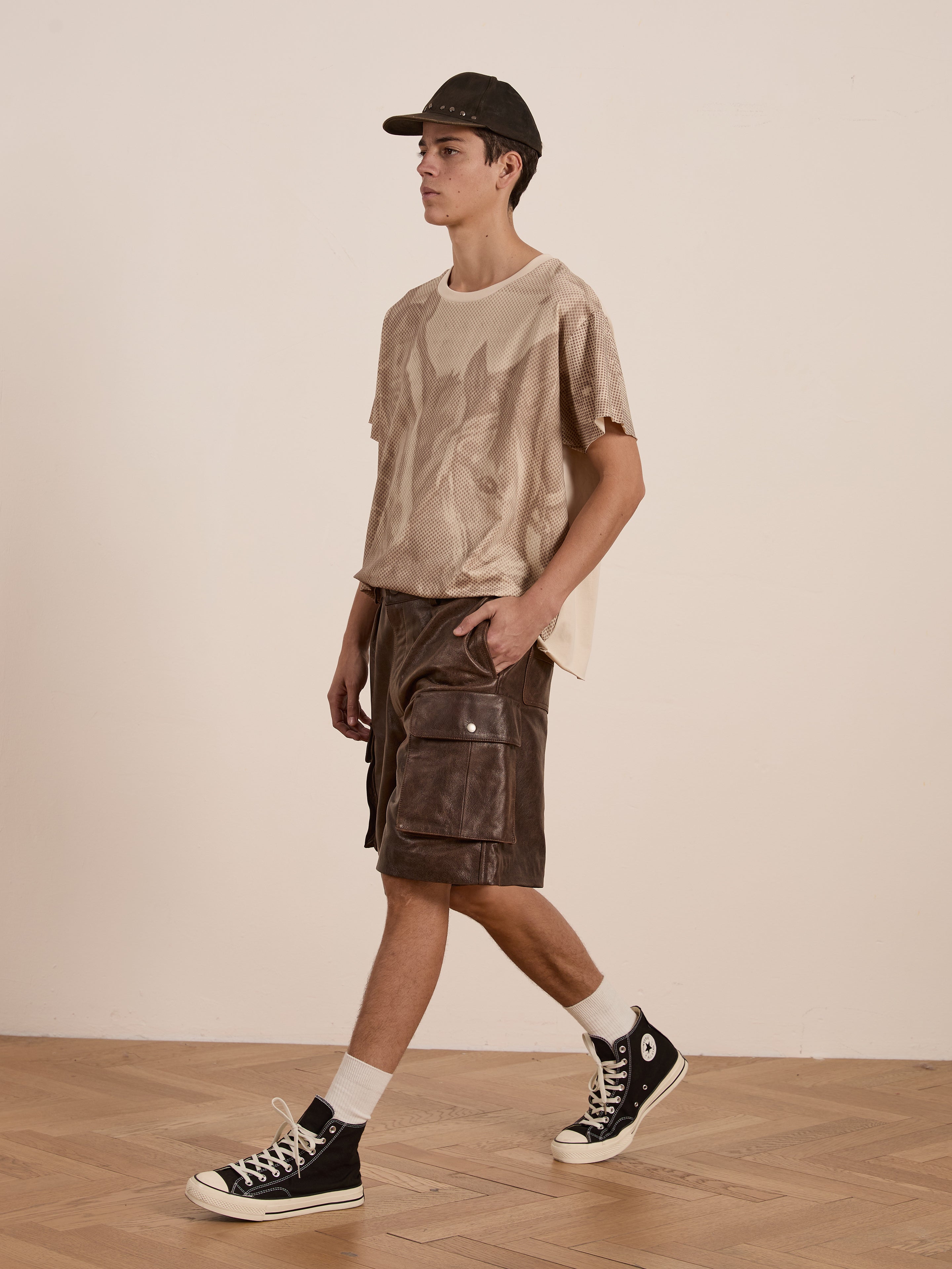 Dressed in a beige FOUND Horse Racer Tee featuring an animal print, brown cargo shorts, black cap, and black sneakers, a person walks on a wooden floor with a relaxed fit.