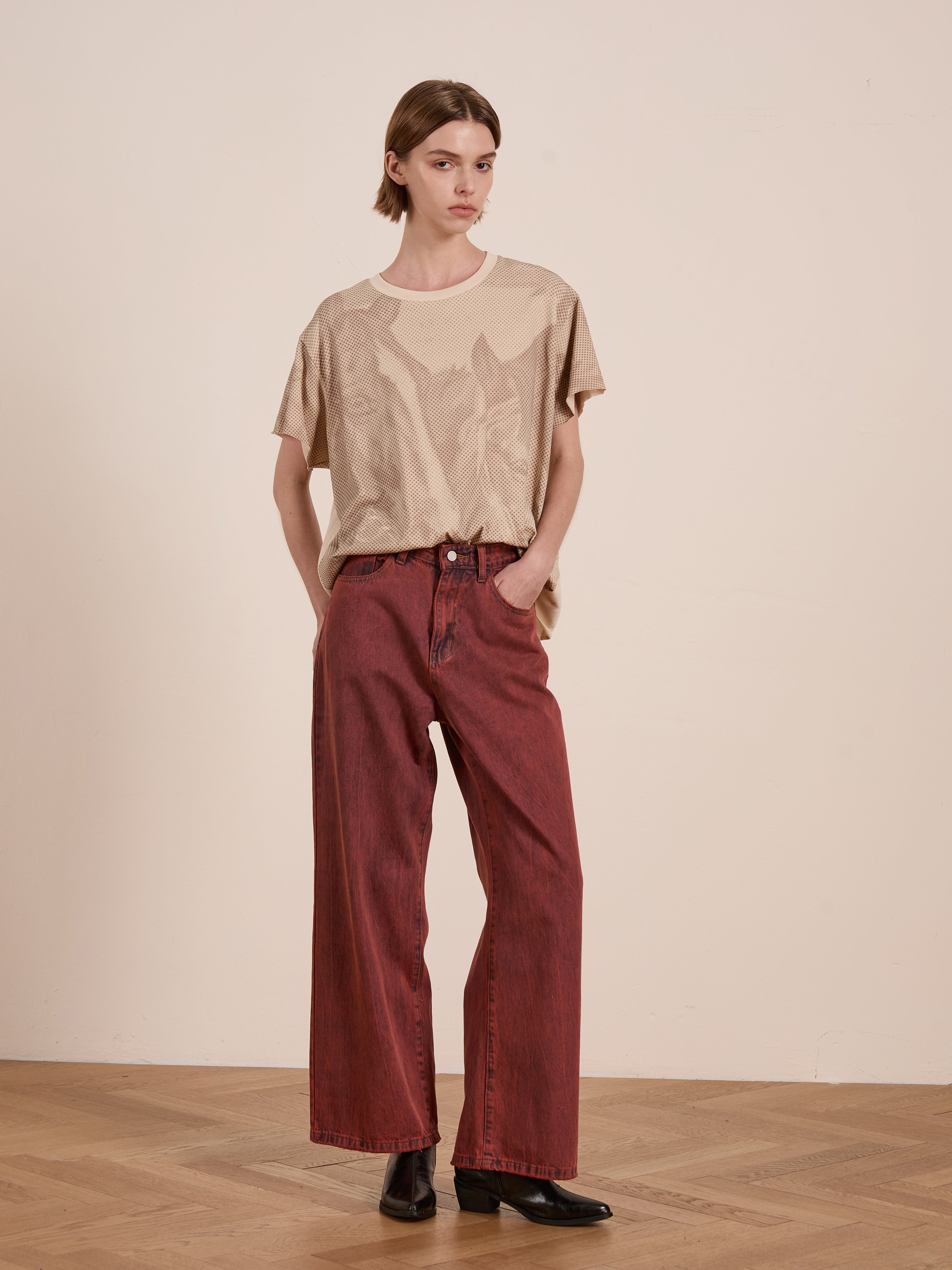 In a wood-floored room, a person models the FOUND Horse Racer Tee, showcasing its relaxed fit and equestrian-inspired beige color. Paired with red wide-legged pants and black boots, their hands rest casually in pockets.