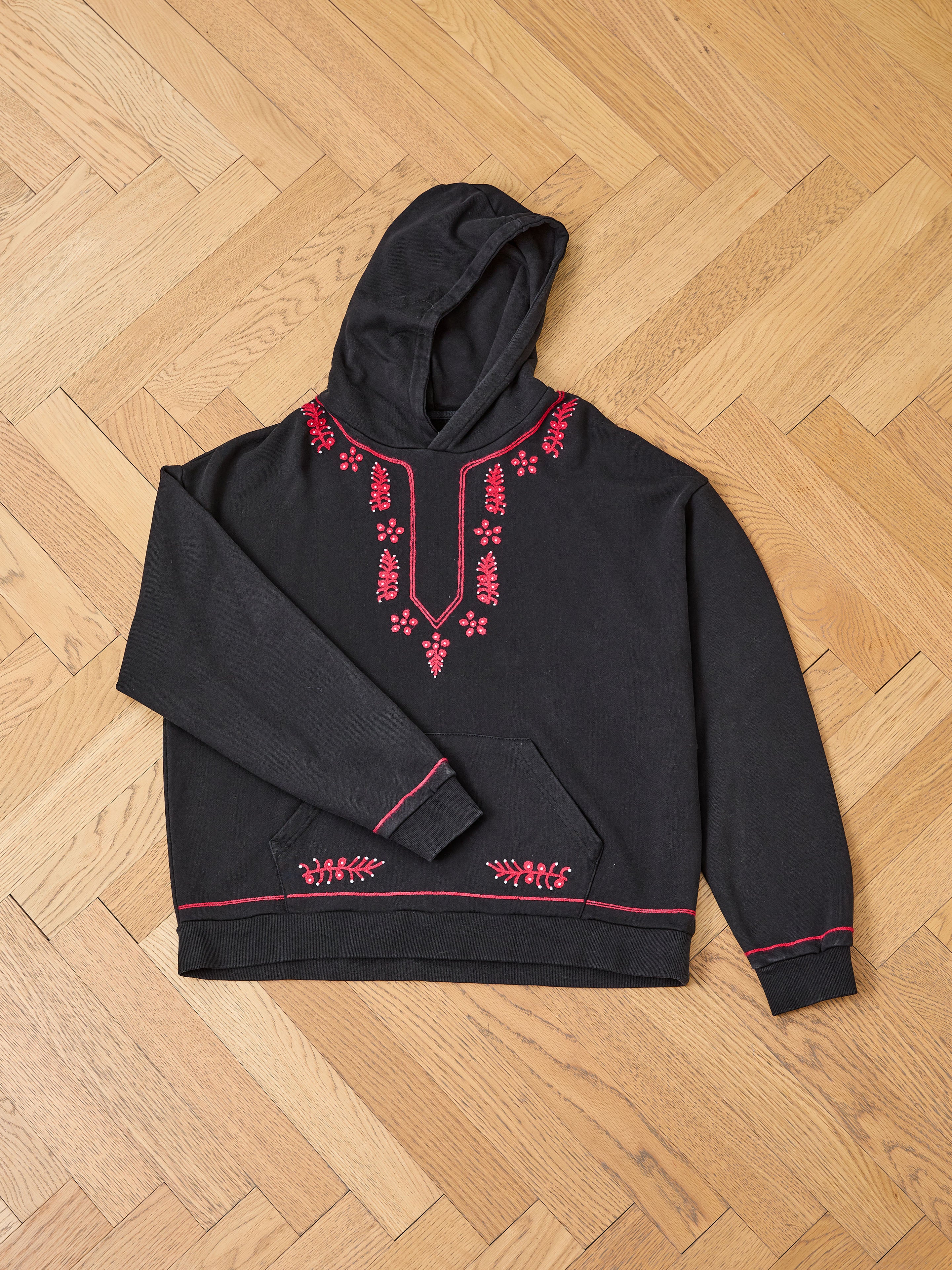 The FOUND Indus Embroidered Hoodie is black with red floral embroidery on the neckline, shoulders, pocket, and sleeves, shown laid flat on a wooden floor.
