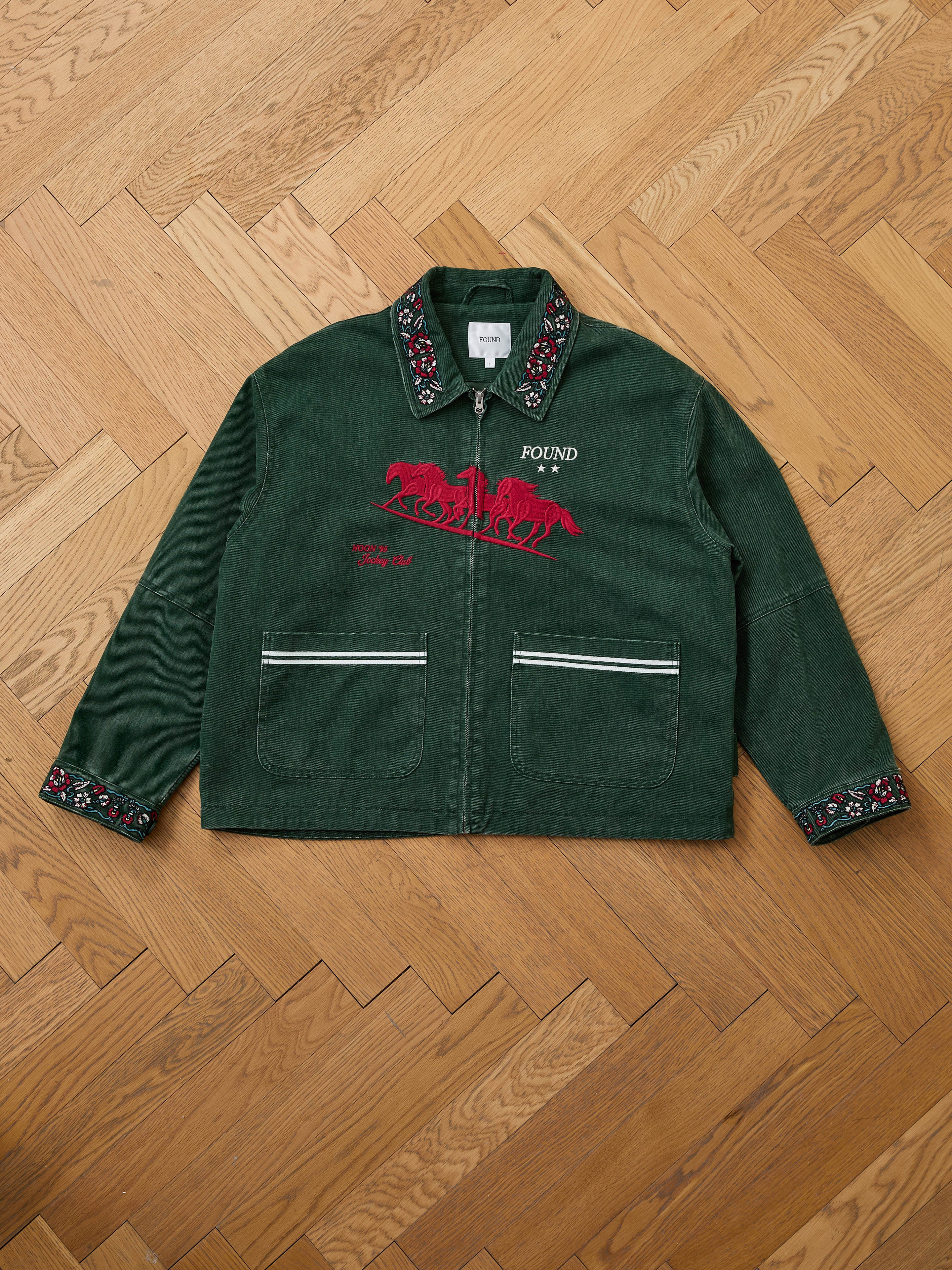 The Found Jockey Club Embroidered Jacket is a green unisex relaxed fit piece featuring racing horses on the front, floral collar and cuffs, two front pockets, and is shown laid flat on a wooden floor.