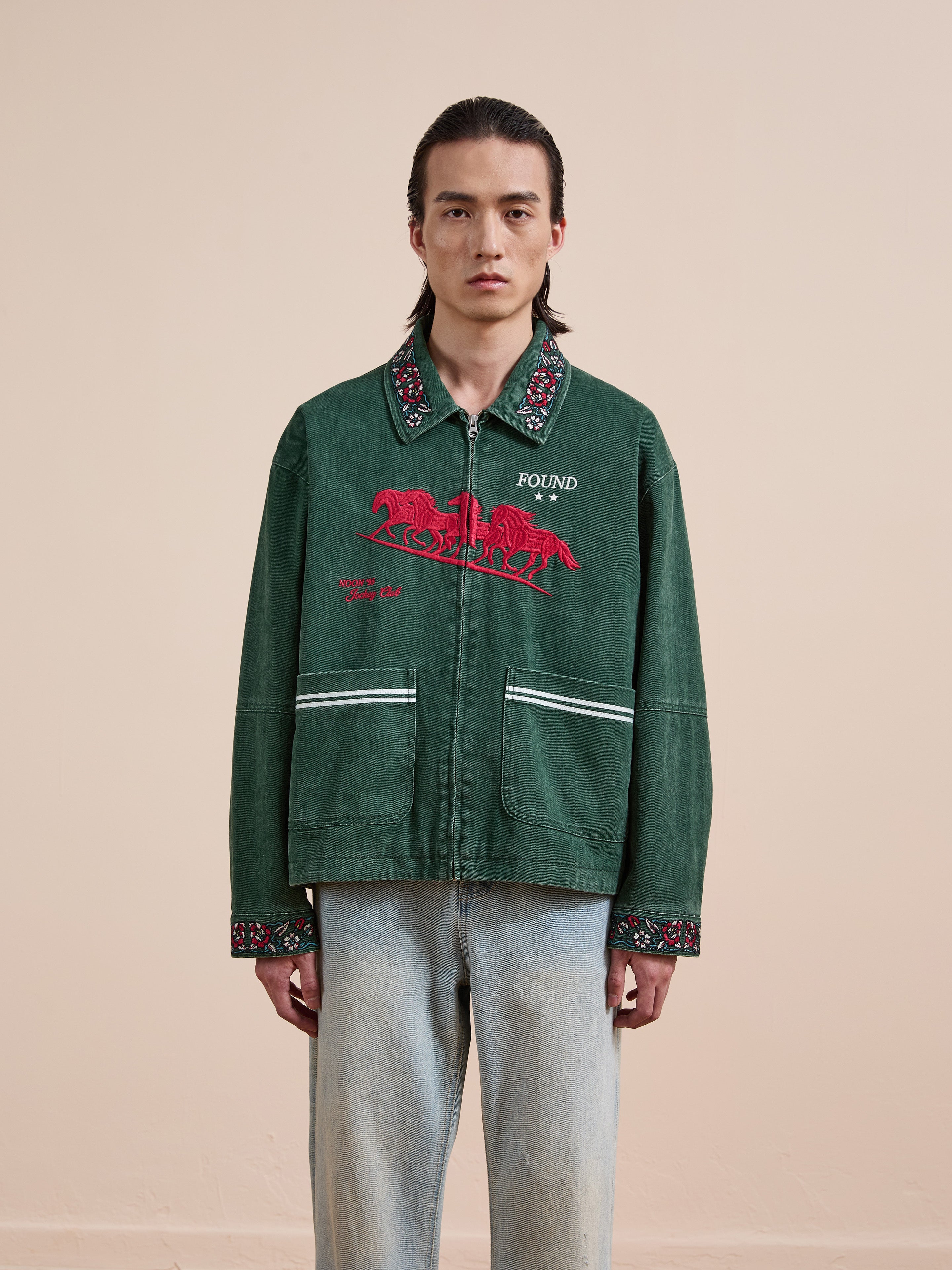 A person with long hair wears the Found Jockey Club Embroidered Jacket and unisex relaxed-fit light blue jeans, standing against a beige background.