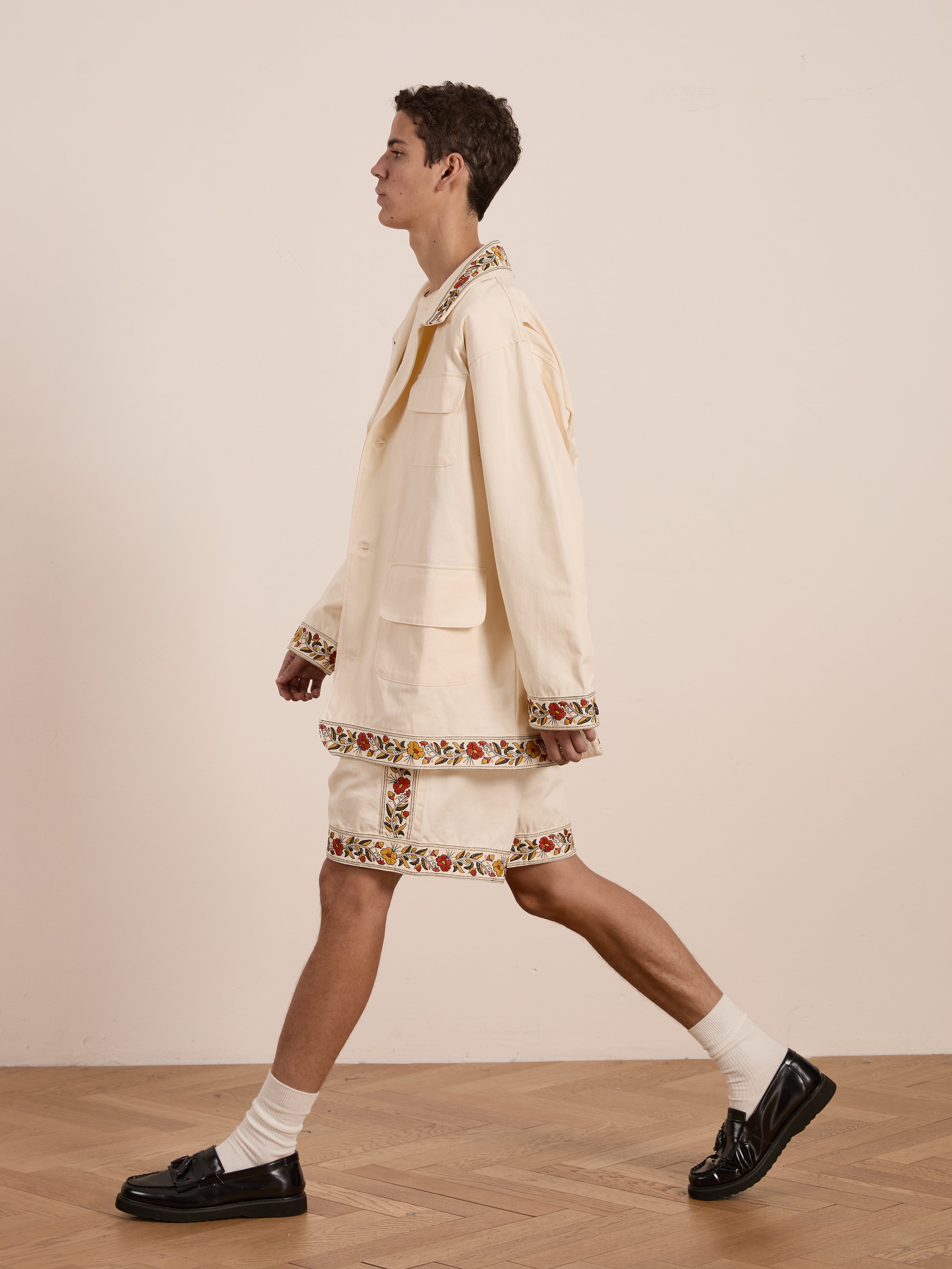 A person walks sideways in a vintage style, wearing FOUND's Kantha Floral Work Jacket with matching light-colored shorts, complemented by white socks and black shoes. The ensemble stands out against a plain background on a wooden floor.