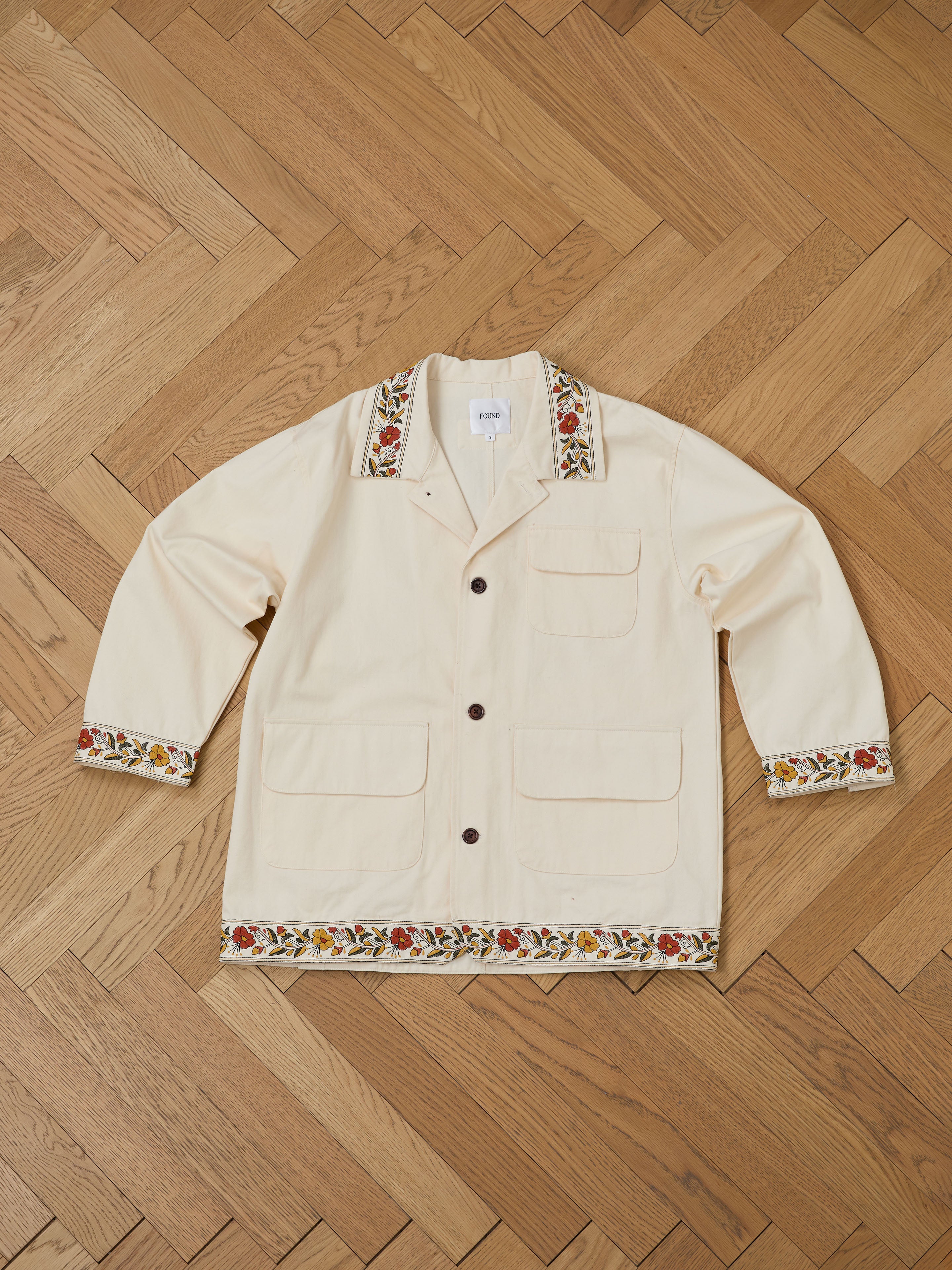 The FOUND Kantha Floral Work Jacket in beige, with intricate embroidery on the cuffs and hem, lies elegantly on a wooden parquet floor, exuding vintage character.