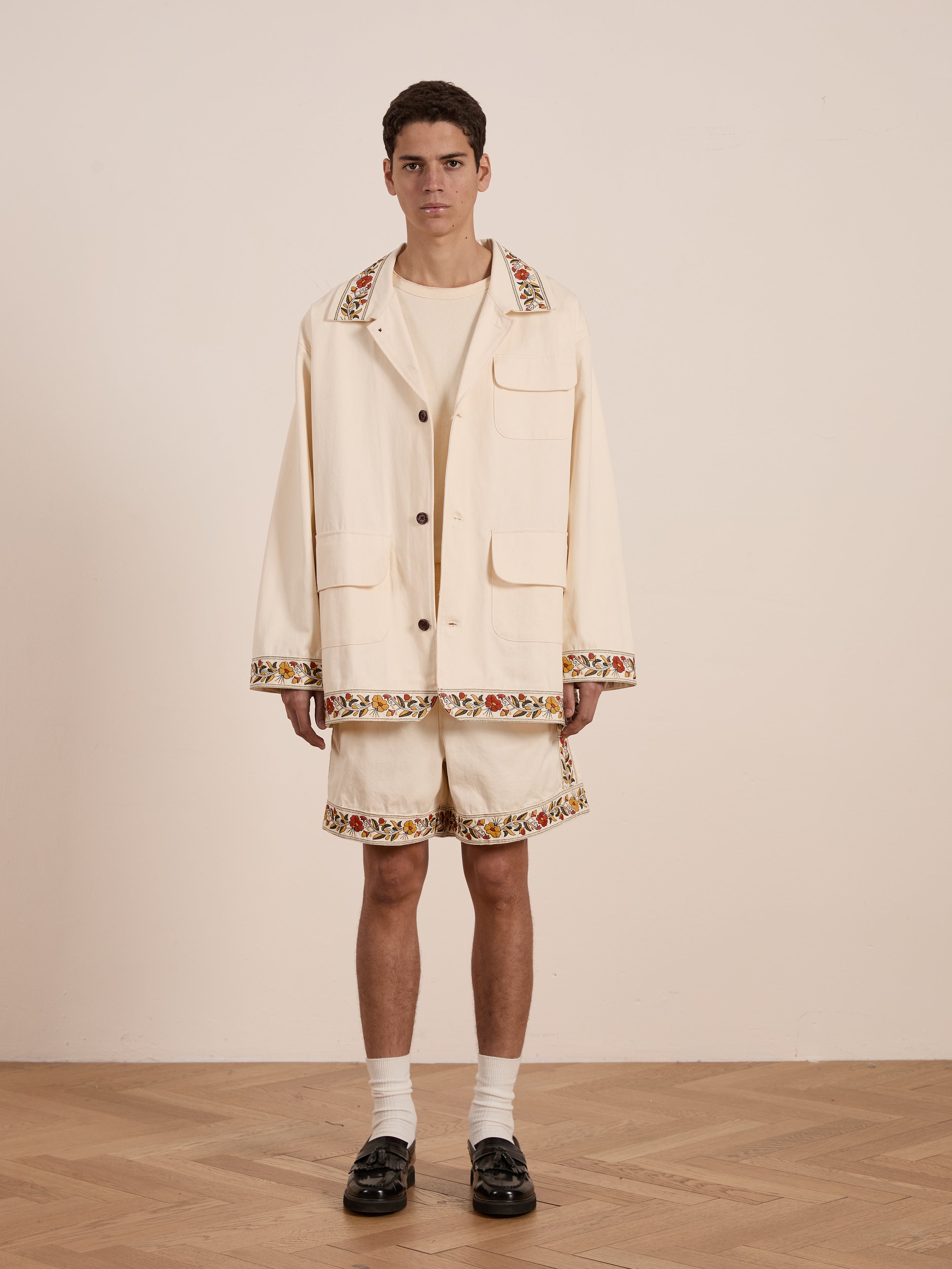 A person stands on a wooden floor wearing the FOUND Kantha Floral Work Jacket, featuring intricate floral embroidery on the collar and hems, exuding a vintage character.