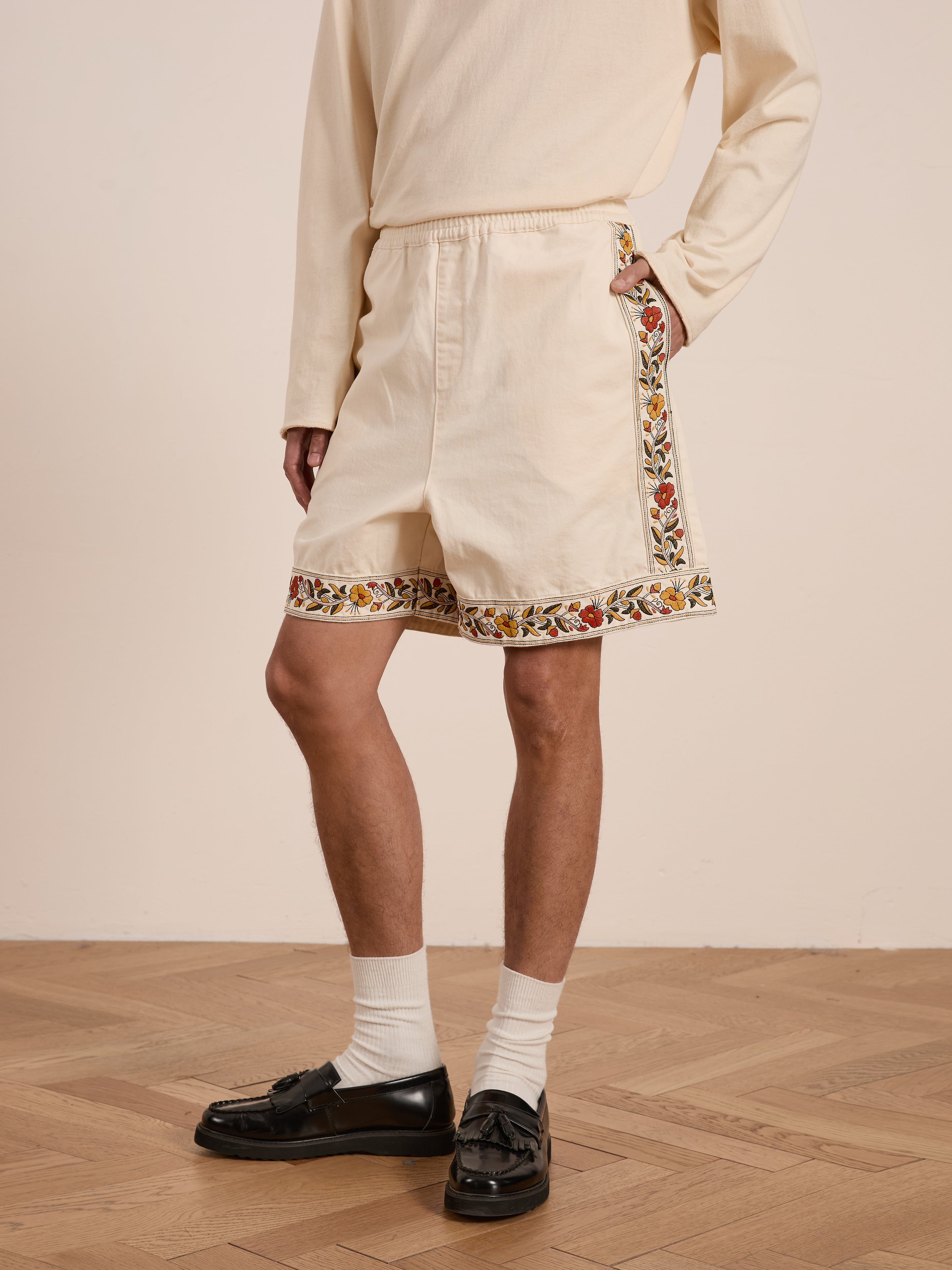 A person in a beige outfit with FOUND's Kantha Floral Work Shorts stands on a wooden floor. These 100% cotton shorts showcase vibrant floral embroidery on the sides and bottom, enhancing the ensemble.