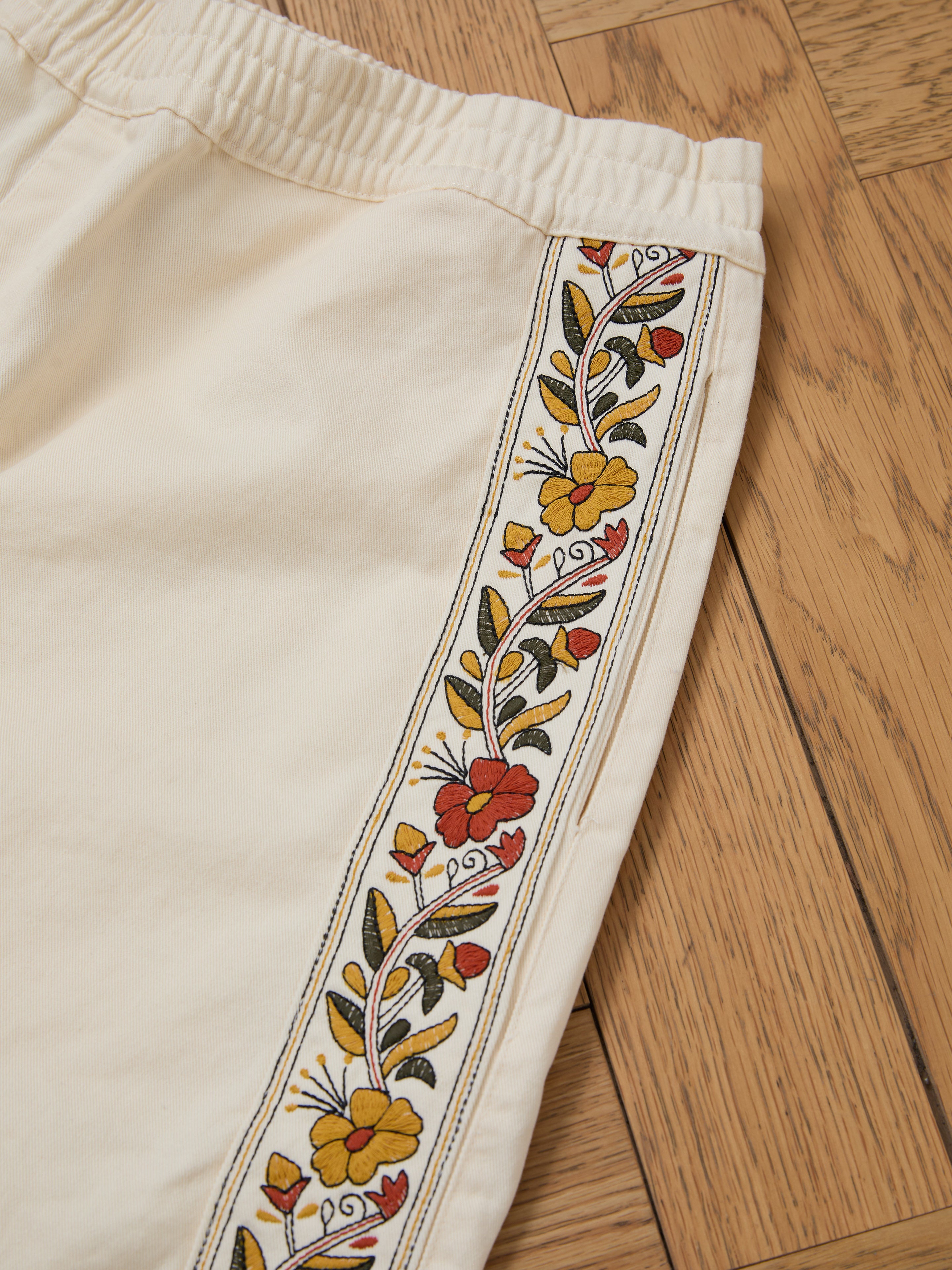 A close-up shot of the Kantha Floral Work Shorts by FOUND, crafted from 100% cotton in beige, highlights exquisite red, yellow, and green floral embroidery along the side. The shorts are elegantly displayed on a wooden floor.