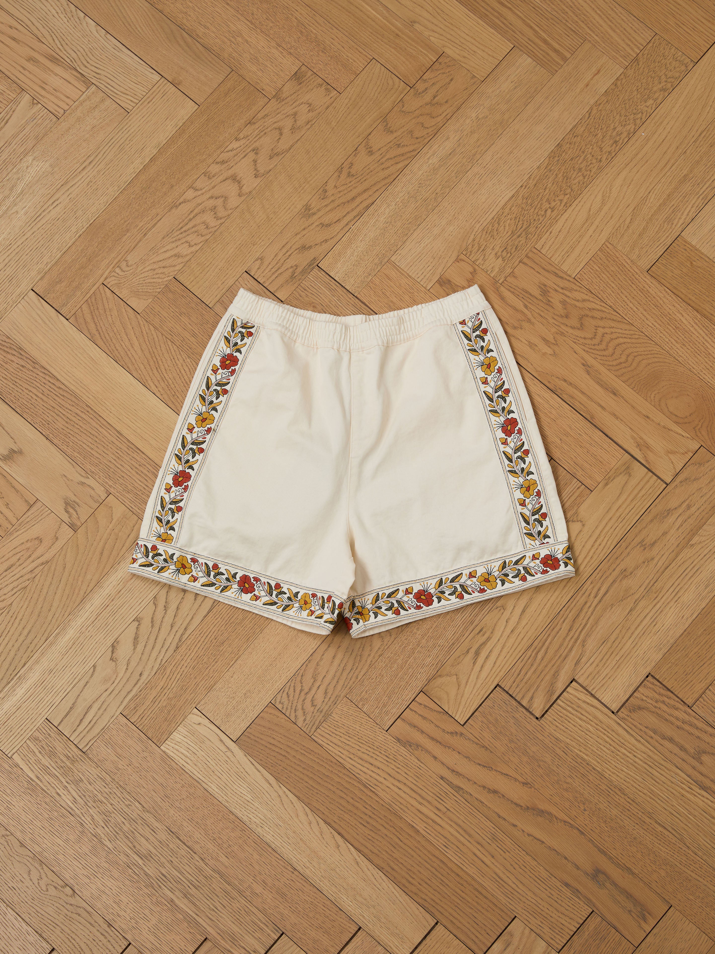 The Kantha Floral Work Shorts by FOUND feature vibrant floral designs along the sides and hem on a neutral cream color, placed on a herringbone wooden floor. Made from 100% cotton, they offer a perfect blend of style and comfort.