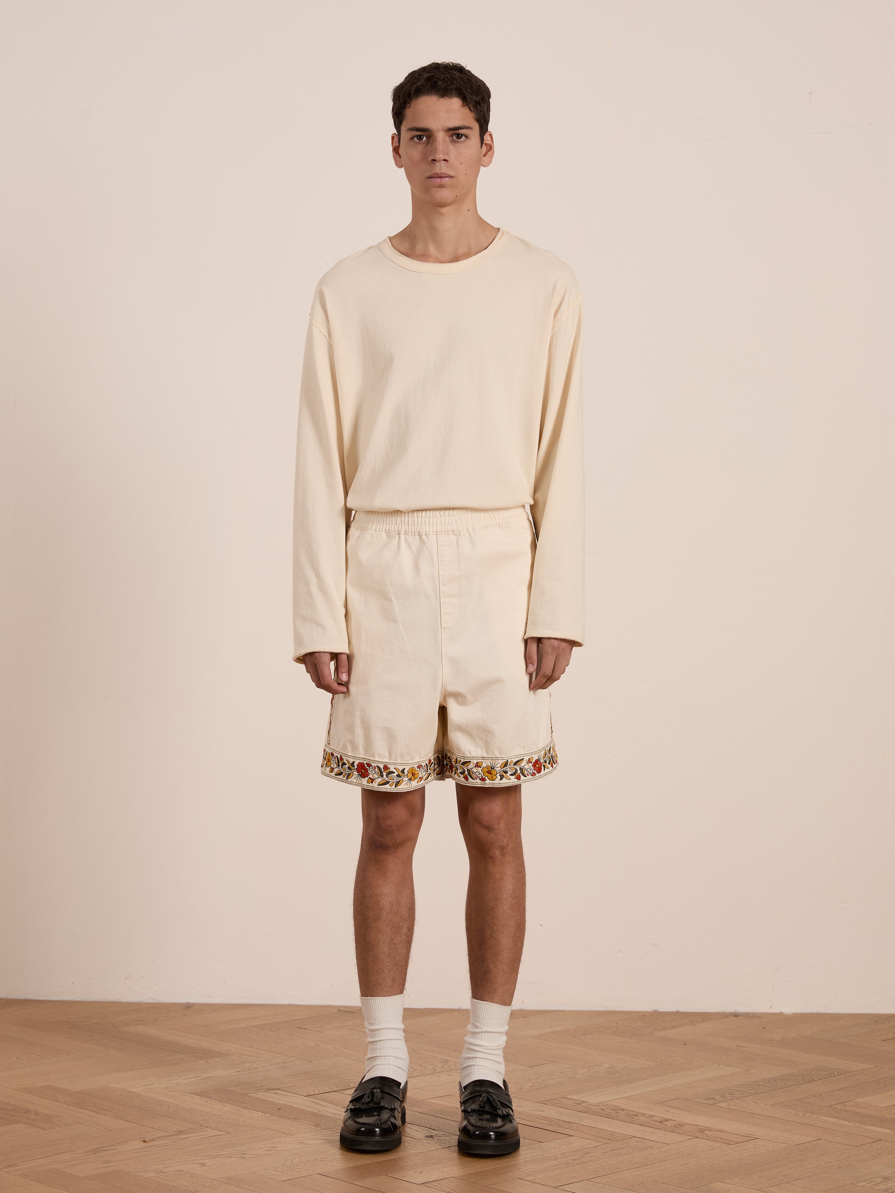 A person stands against a plain background in a cream long-sleeve shirt and FOUND's Kantha Floral Work Shorts featuring exquisite floral embroidery and decorative trim. The look is completed with white socks and dark loafers.