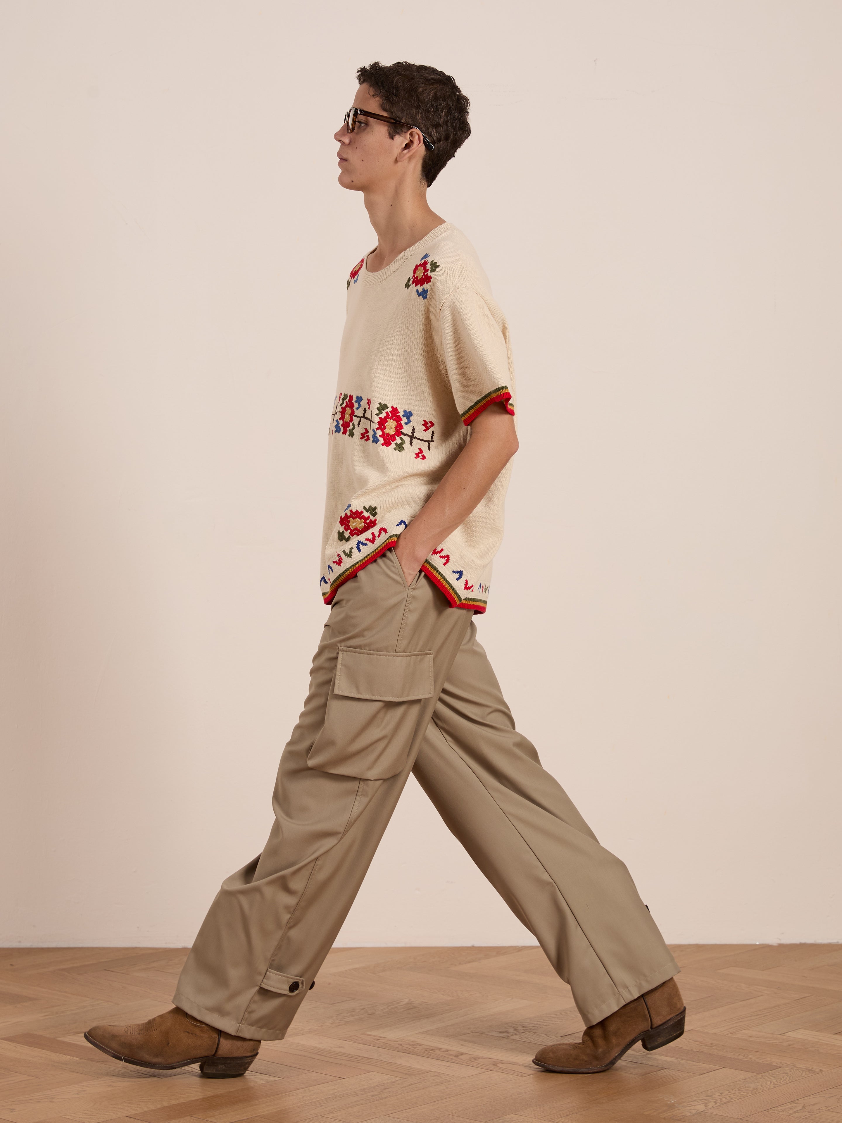 A person strolls on a wooden floor, wearing glasses, the FOUND Knit Flower Petal Tee with vintage-inspired cream and red floral motifs, tan cargo pants, and brown shoes.