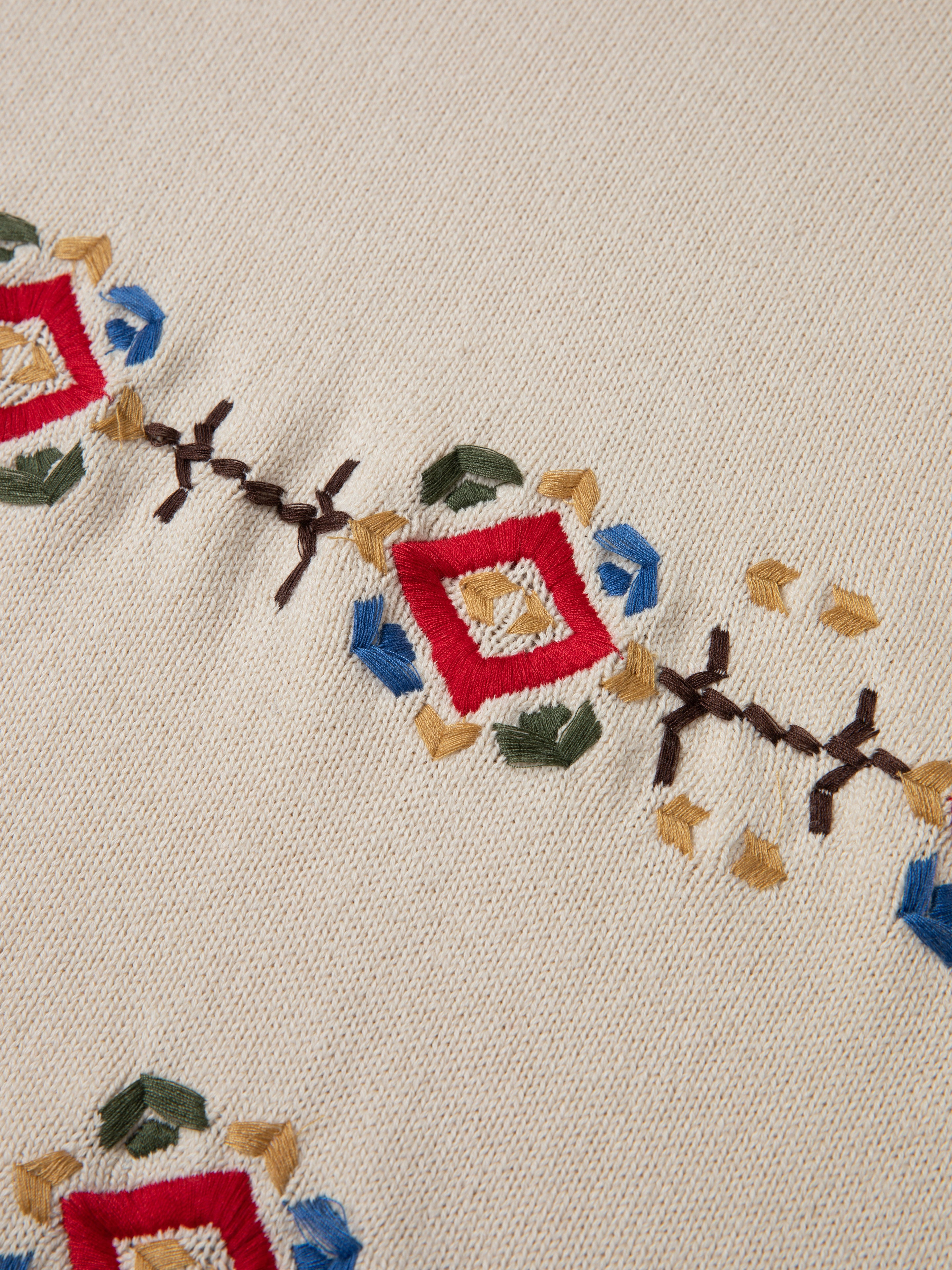 A beige fabric from the Knit Flower Petal Tee by FOUND showcases a vintage-inspired charm with geometric embroidered patterns in red, blue, yellow, and green shapes.