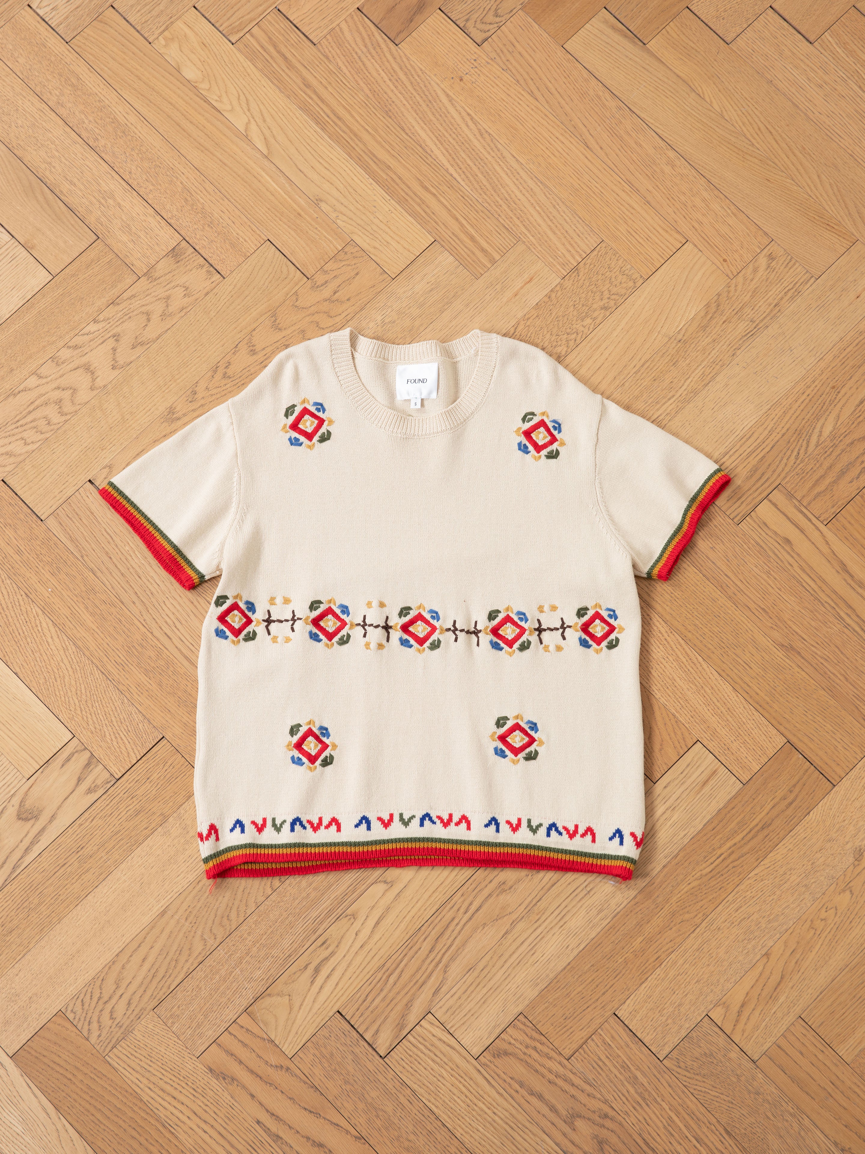 The Knit Flower Petal Tee by FOUND is a beige knit shirt with vintage charm, showcasing colorful geometric embroidery and striped red, army green, and brown trims on the sleeves and hem. Laid on a herringbone wood floor, its design mirrors jacquard floral motifs.