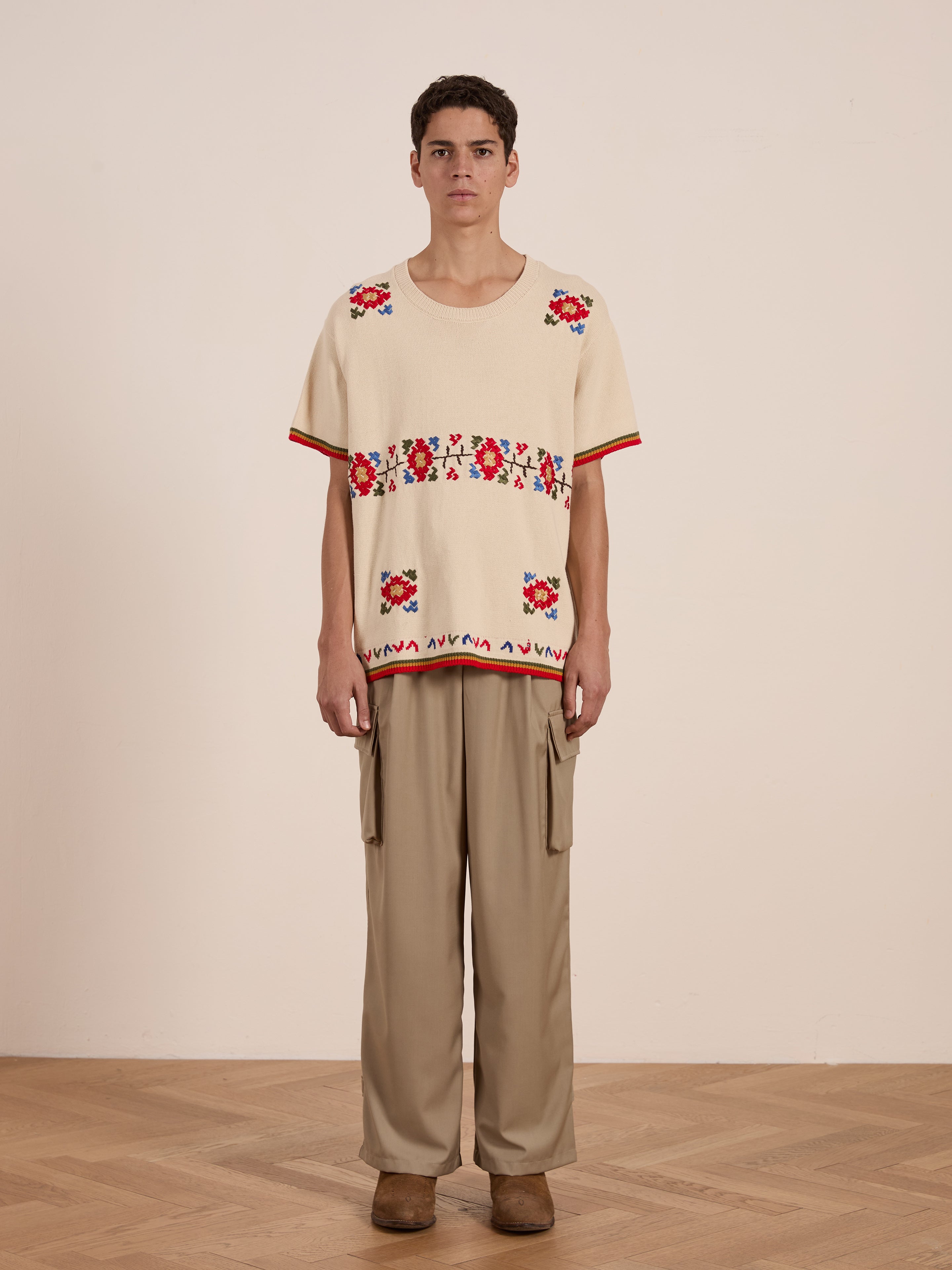 Indoors, an individual models the FOUND Knit Flower Petal Tee with jacquard floral designs, teamed with beige cargo pants and brown shoes, posed before a plain wall and wooden floor.