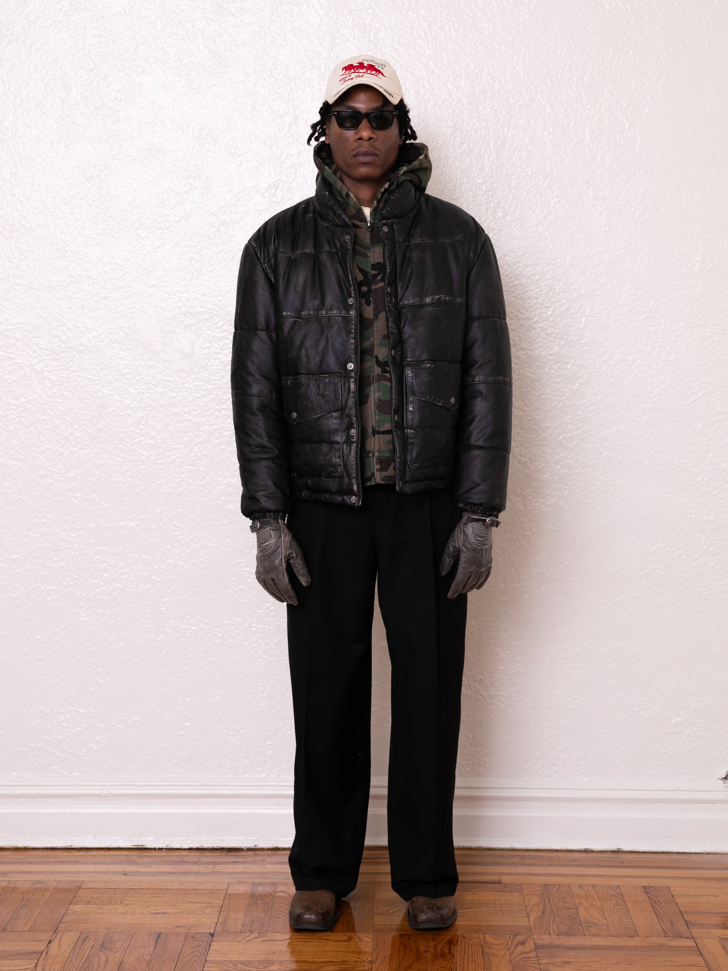 A person in a FOUND Leather Distressed Puffer Jacket wears a camouflage hoodie, black pants, sunglasses, gray gloves, white cap, and brown shoes while standing against a white wall on a wooden floor.