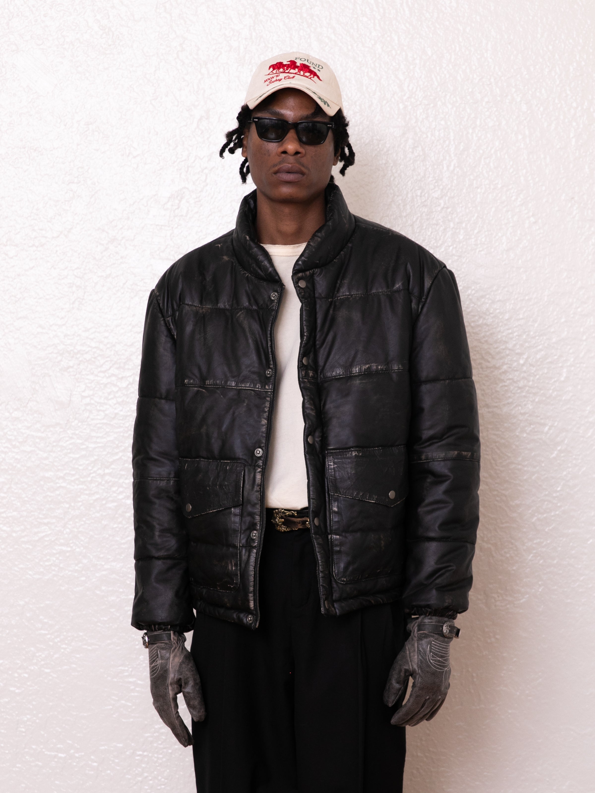 A person wearing a FOUND Leather Distressed Puffer Jacket, white shirt, dark pants, gloves, sunglasses, and a white cap stands against a textured white wall.