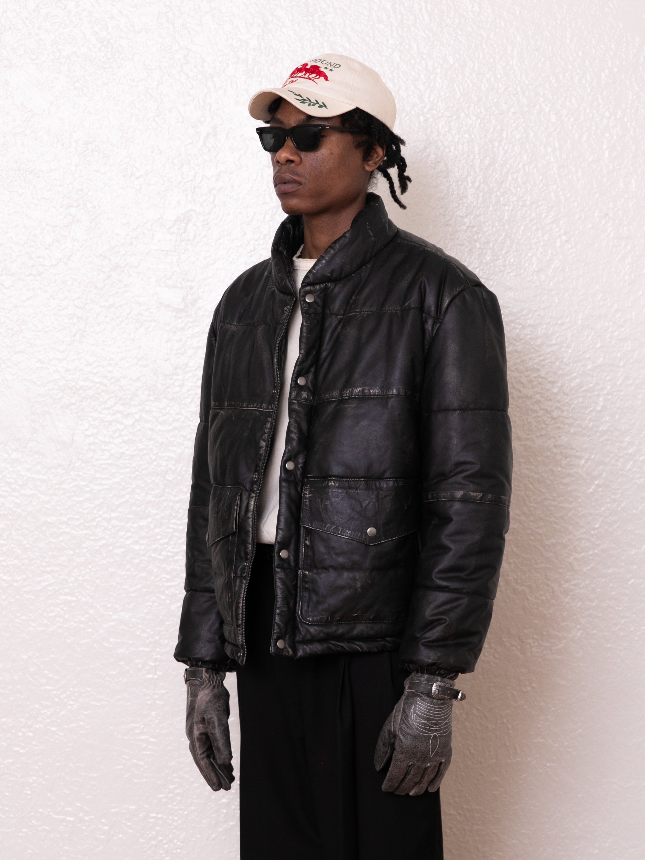 A person in a FOUND Leather Distressed Puffer Jacket, black pants, sunglasses, gray gloves, and a white baseball cap with red lettering stands against a textured white wall.