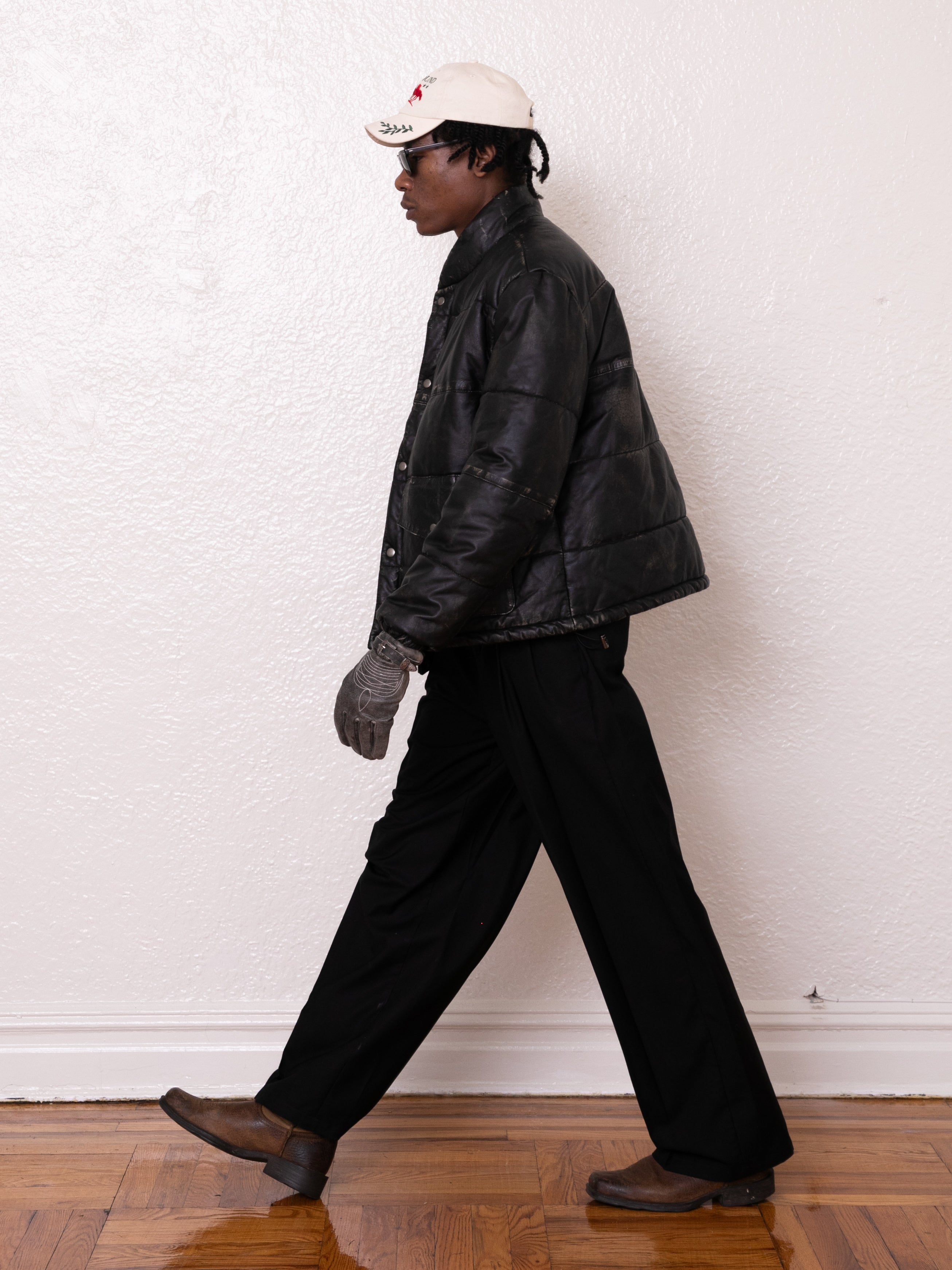 A person walks sideways in a black FOUND Leather Distressed Puffer Jacket, black pants, and brown shoes. They wear a white cap and gloves against a white wall. The ethically sourced jacket adds eco-friendly flair to their look.