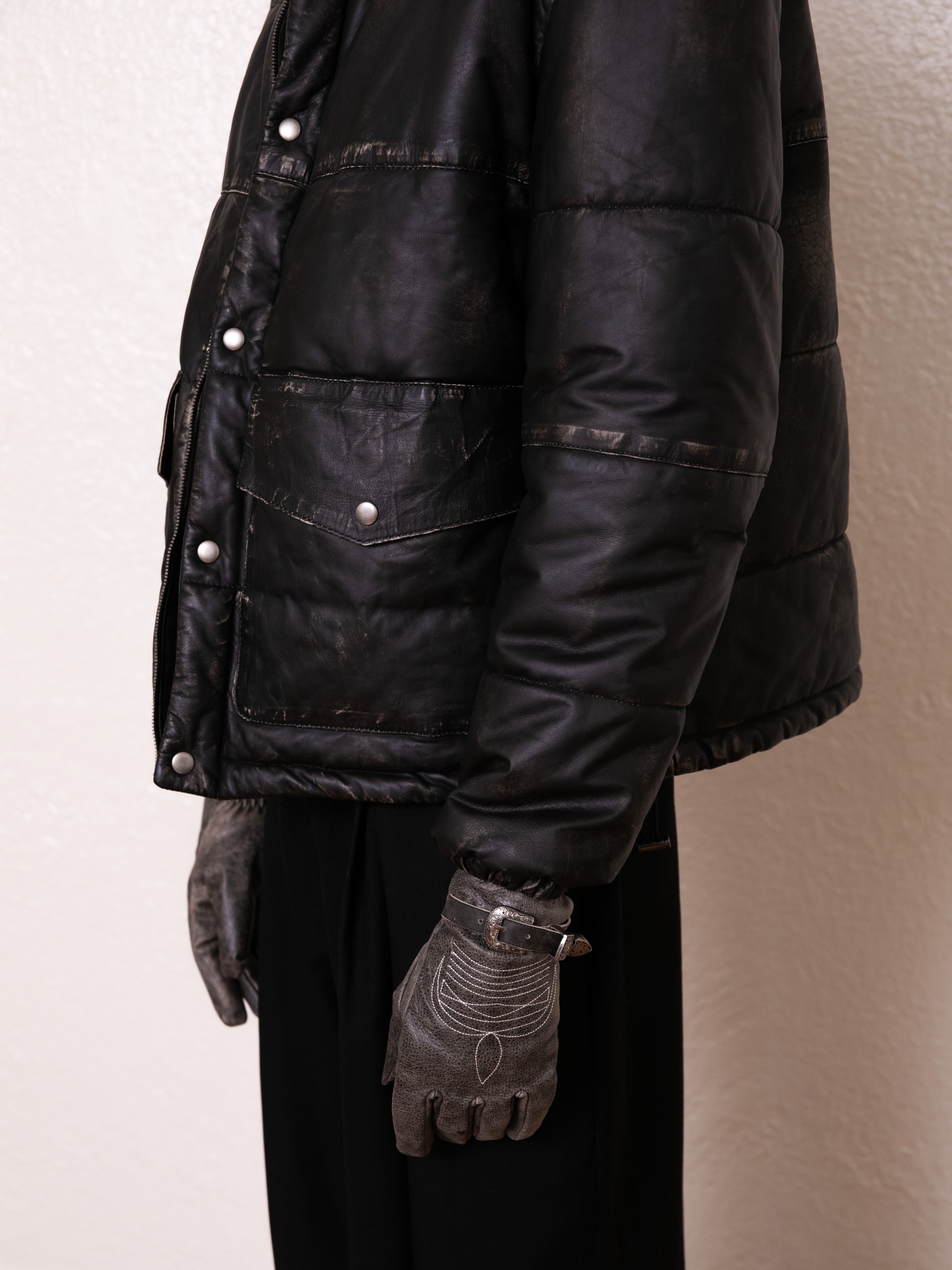 A person models the FOUND Leather Distressed Puffer Jacket with gray gloves, standing sideways against a plain background, exemplifying a stylish and sustainable choice made from ethically sourced materials.