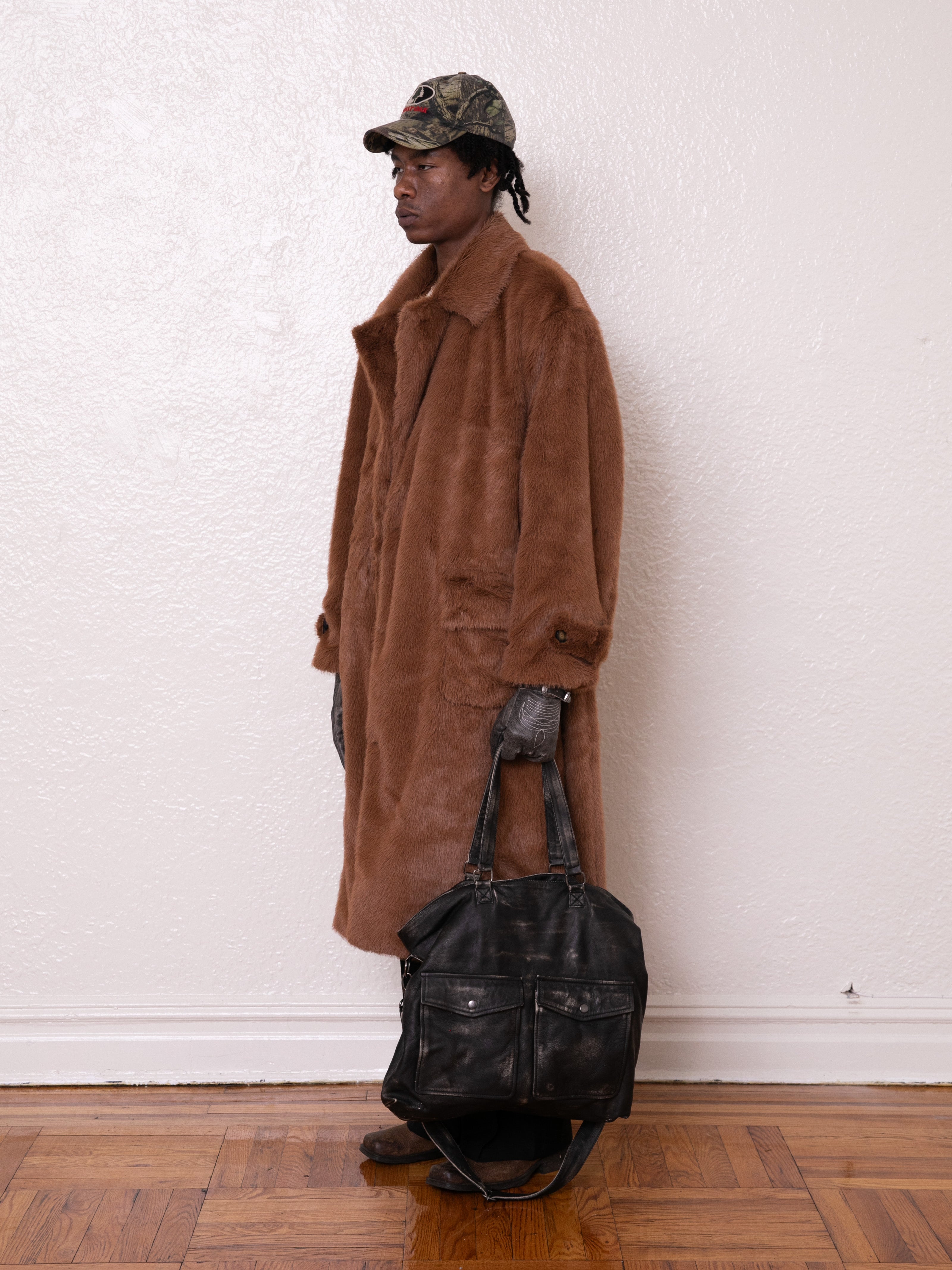 A person is standing against a plain wall, wearing FOUND's Long Faux Fur Coat and a camo cap while holding a sleek black leather bag.