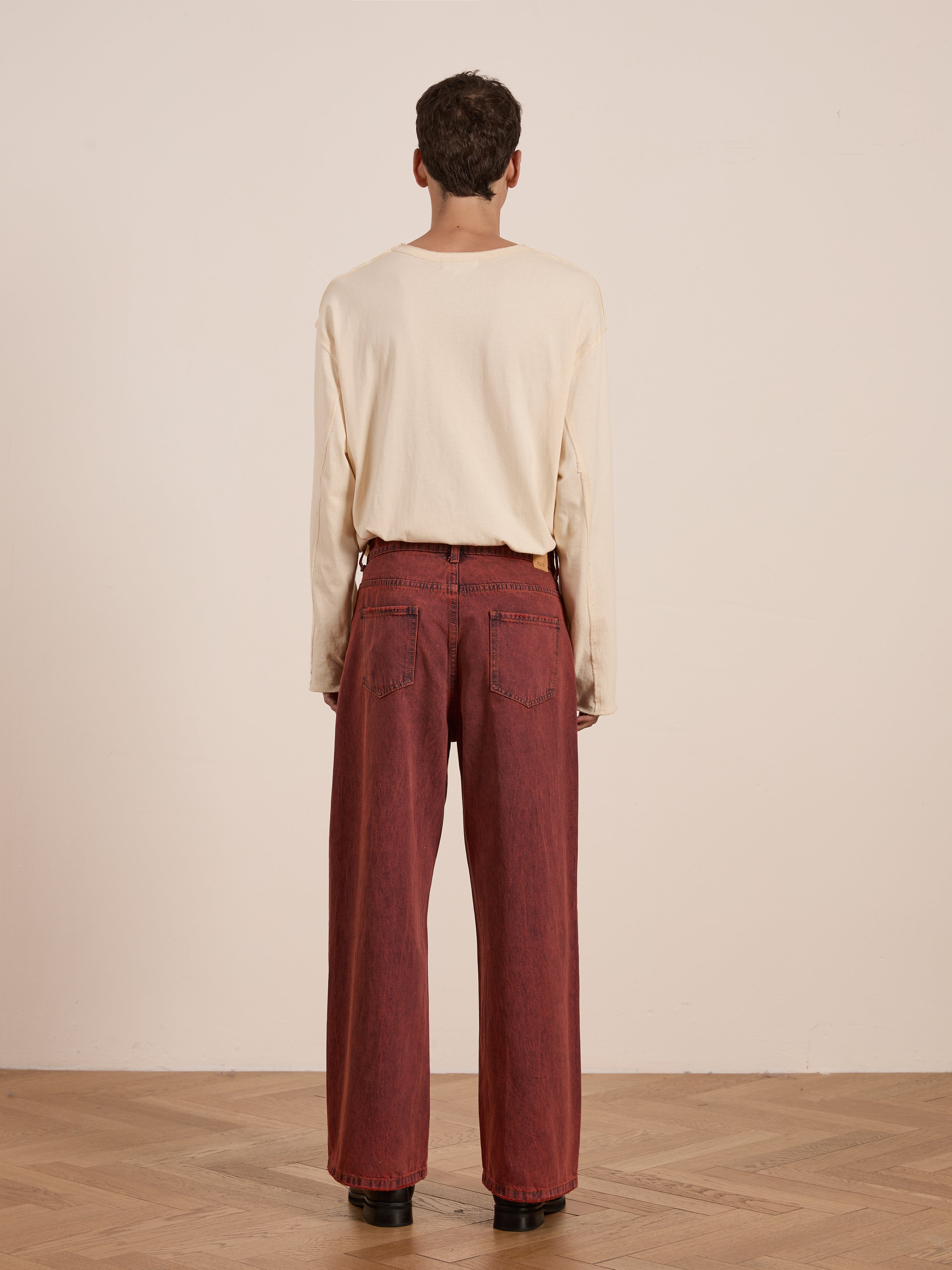 A person in vintage red wash, beige long-sleeve shirt and FOUND's Lacy Baggy Jeans stands facing a plain background, with a wooden floor beneath.