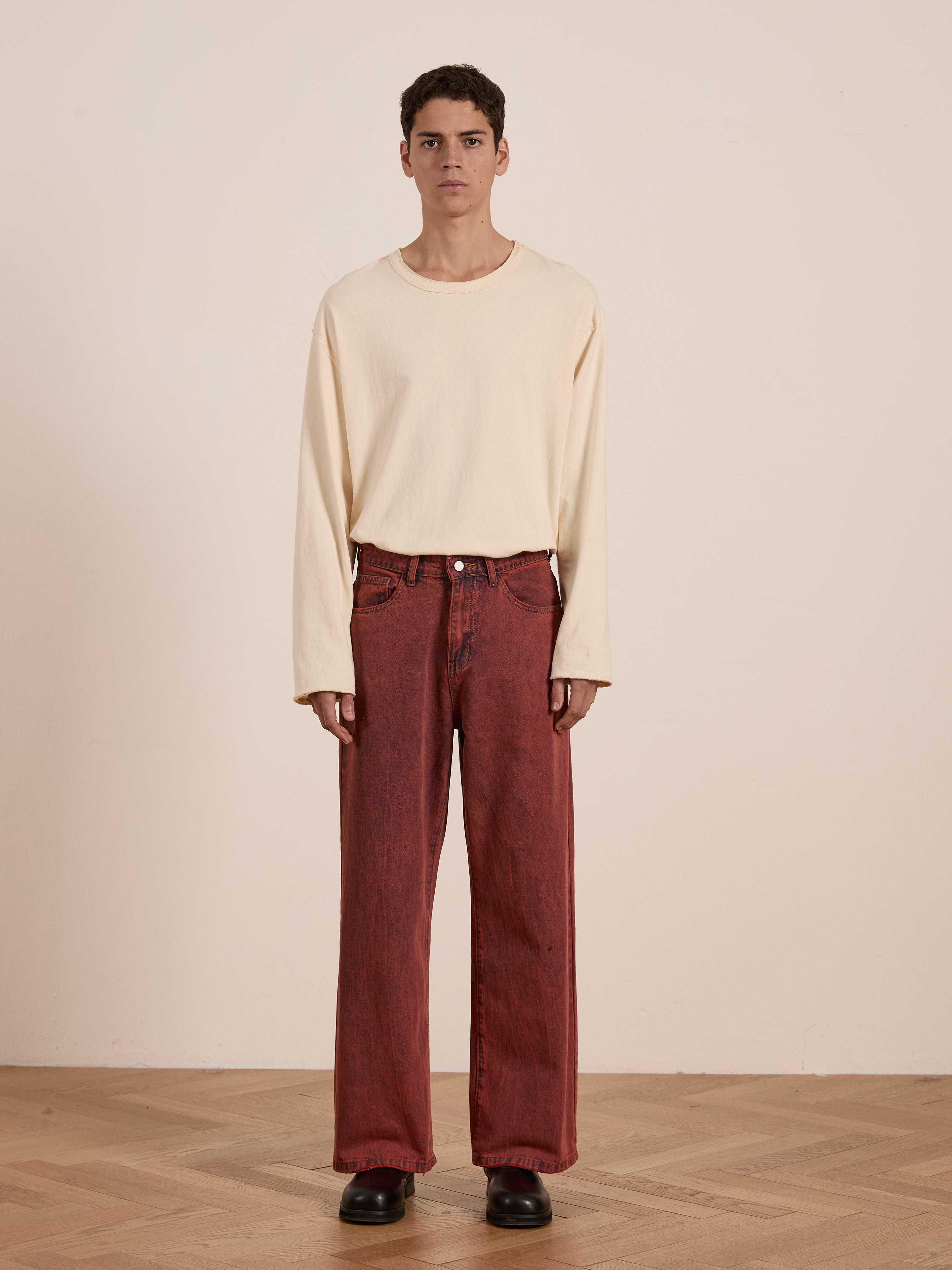 Indoors, a person wears a cream long-sleeve shirt with FOUND's Lacy Baggy Jeans in vintage red wash, made from 100% cotton, paired with black shoes on a wooden floor.