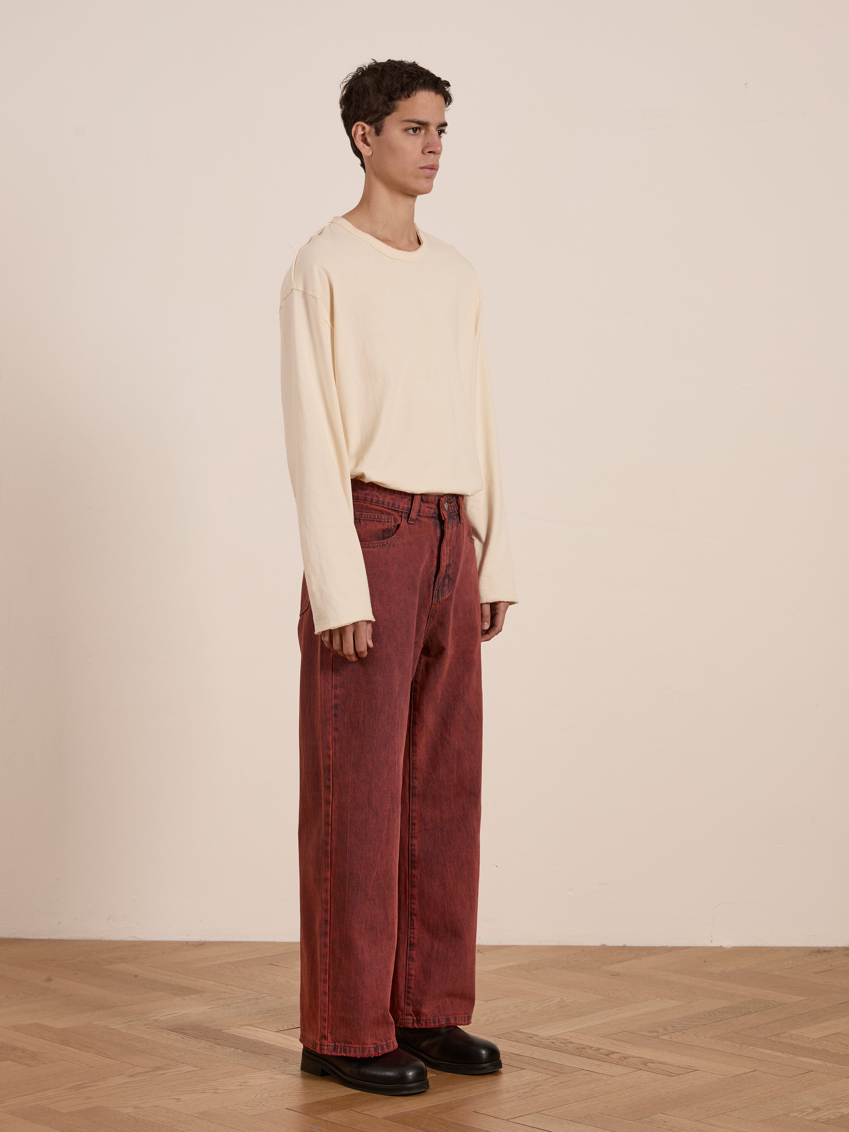 A person wearing a beige long-sleeve shirt, FOUND's Lacy Baggy Jeans in vintage red wash, and black shoes stands on a wooden floor against a plain wall.