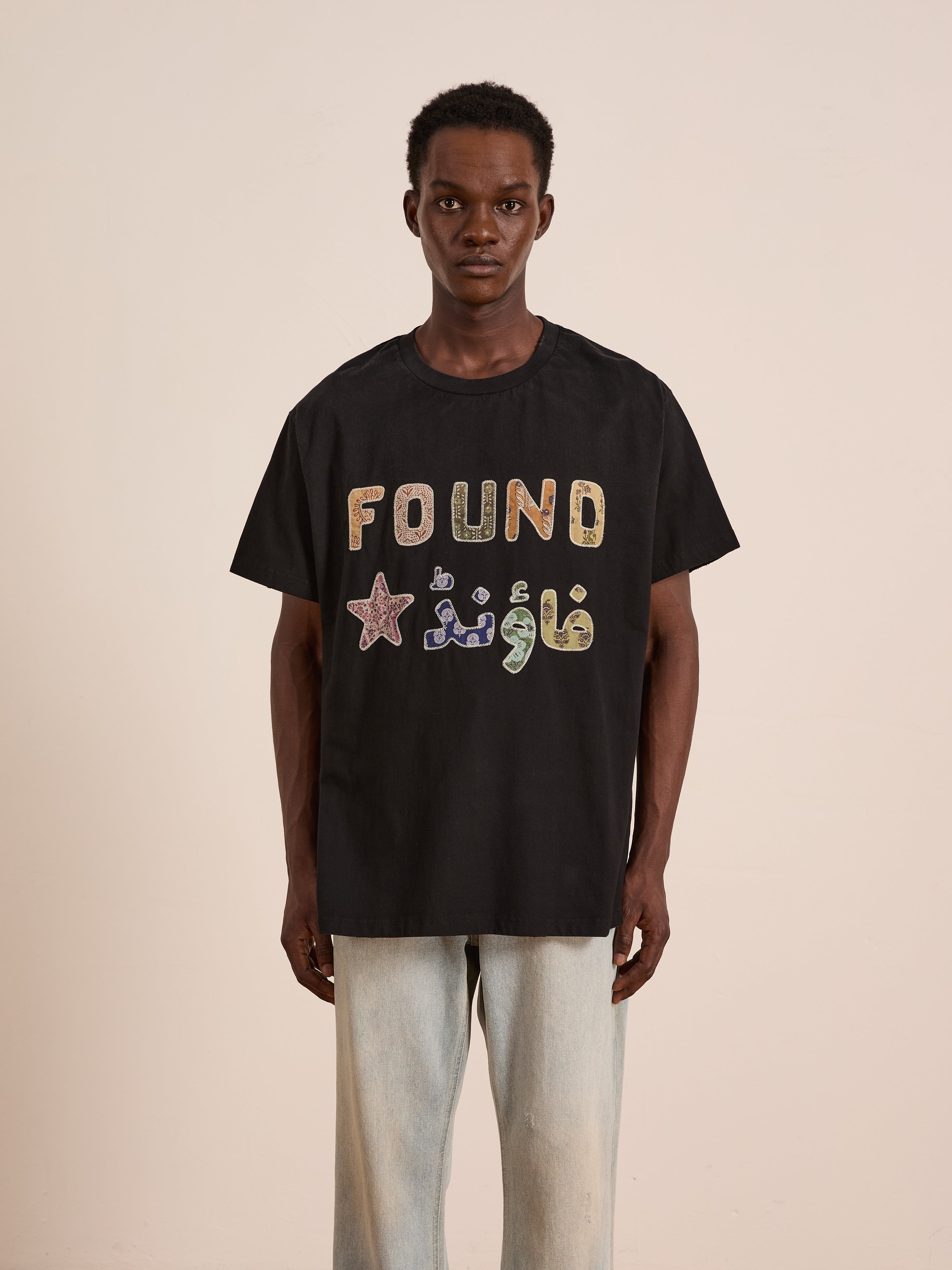 A person faces forward wearing the FOUND Language Tapestry Logo Tee—a black shirt with "FOUND" and Arabic text in colorful letters, a vintage-inspired fade, and star graphic—paired with light-colored pants.