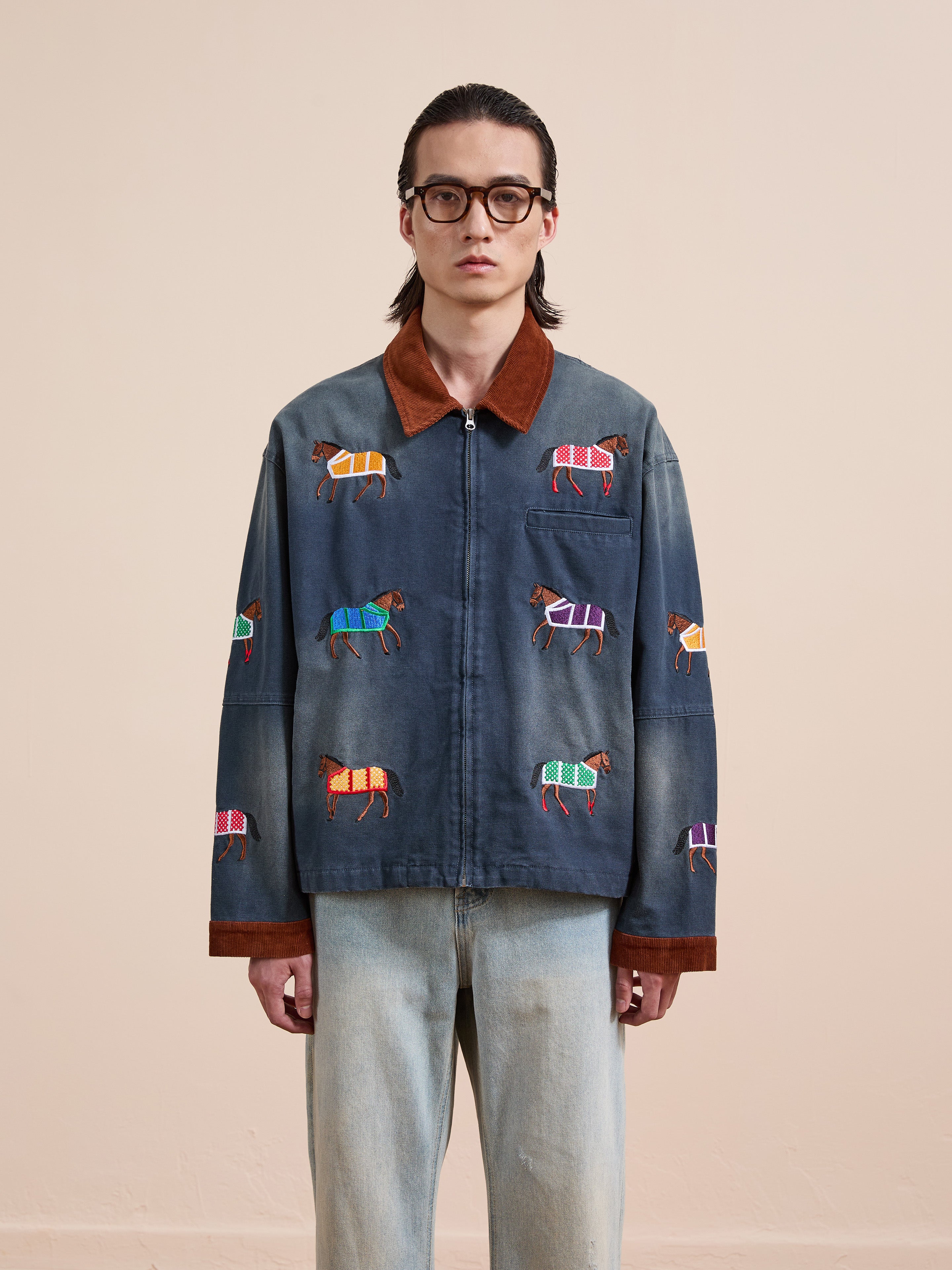 A person wearing glasses models the Found Lariat Horse Embroidered Work Jacket, featuring colorful horses, while facing the camera against a plain beige background.