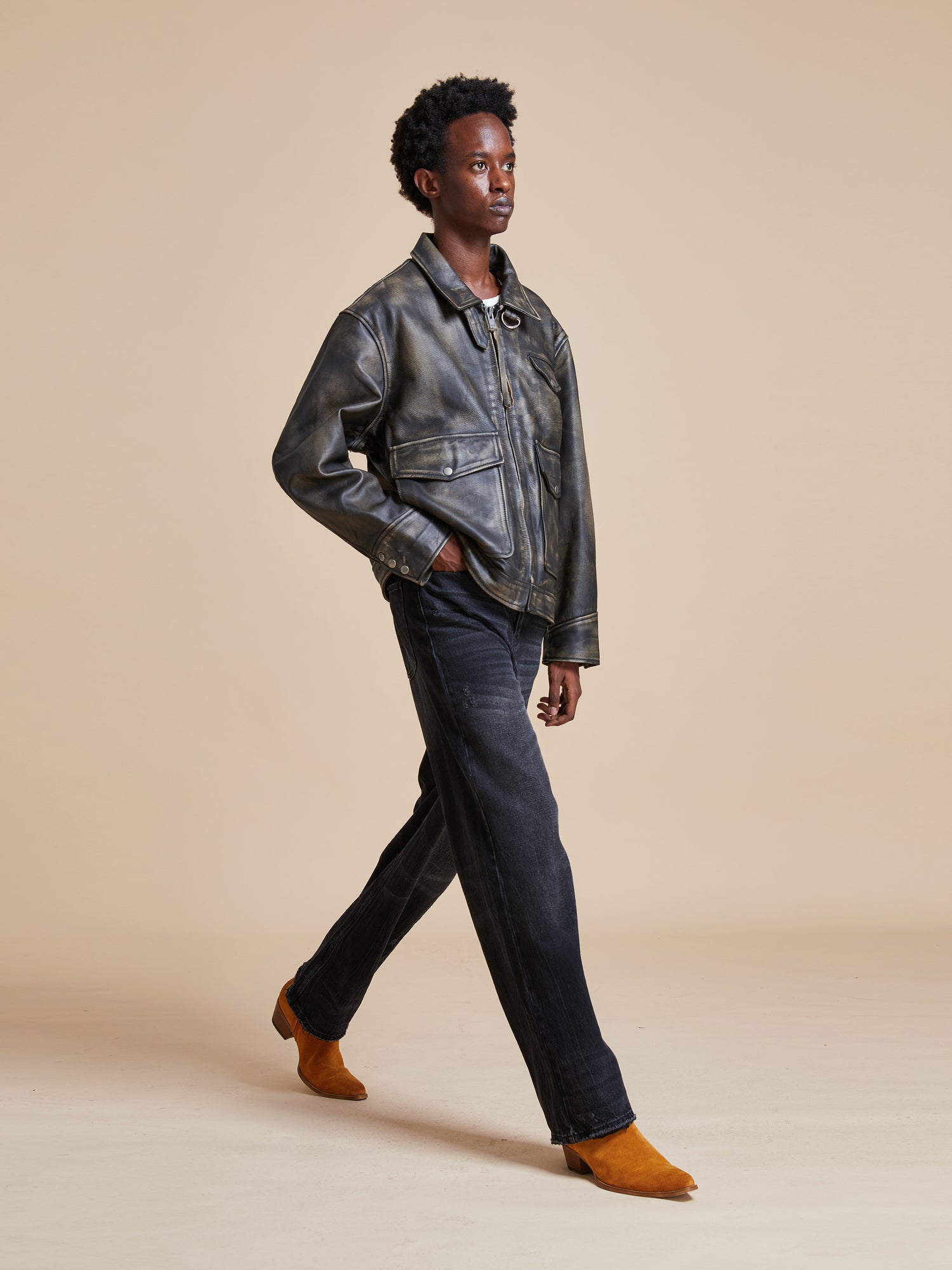 A man in a Found distressed leather pocket jacket and jeans walking on a beige background.