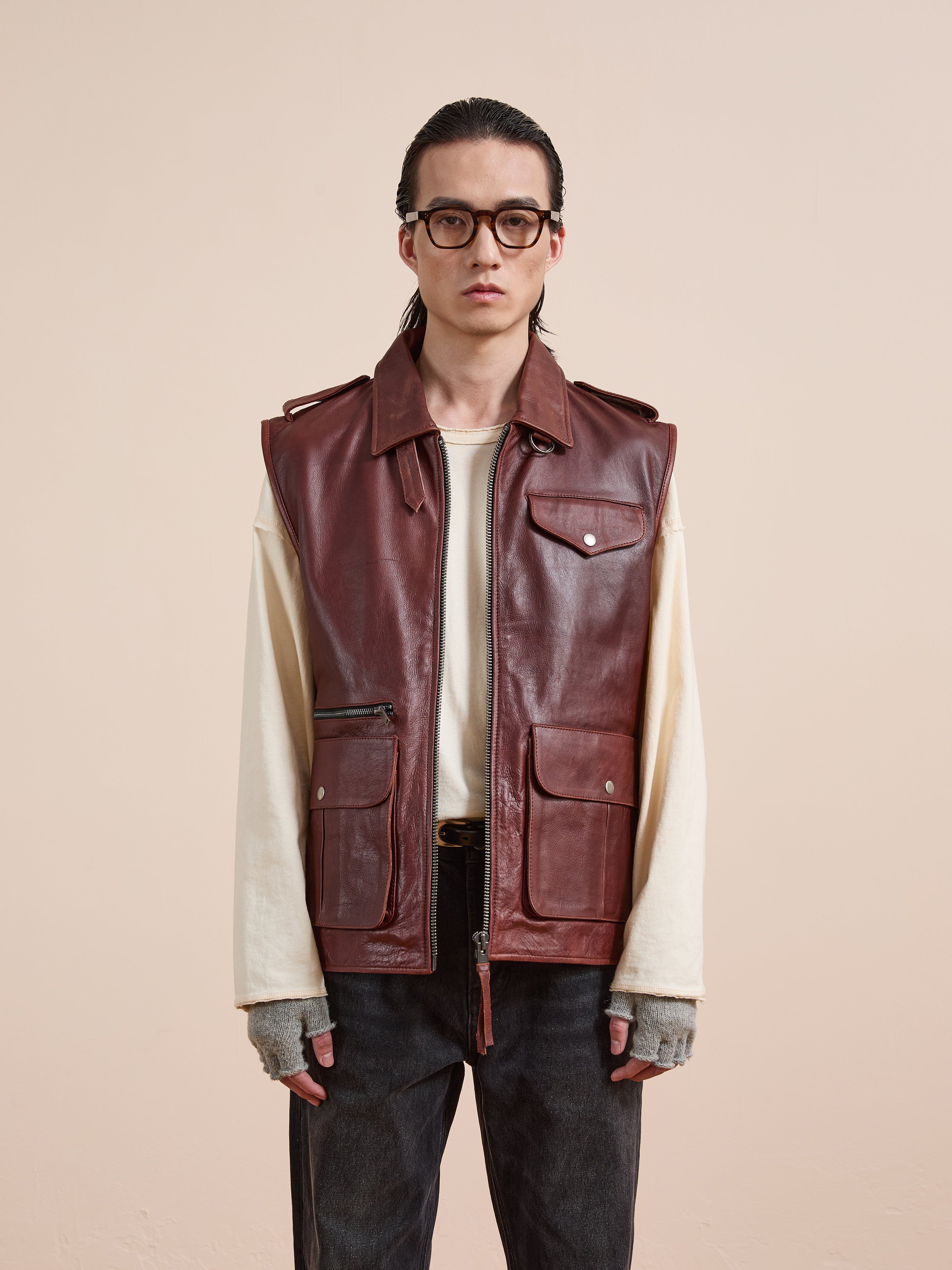 A person wearing glasses and Found's distressed brown Leather Pocket Vest with utility pockets over a cream long-sleeve shirt, gray fingerless gloves, and dark jeans stands against a plain beige background.