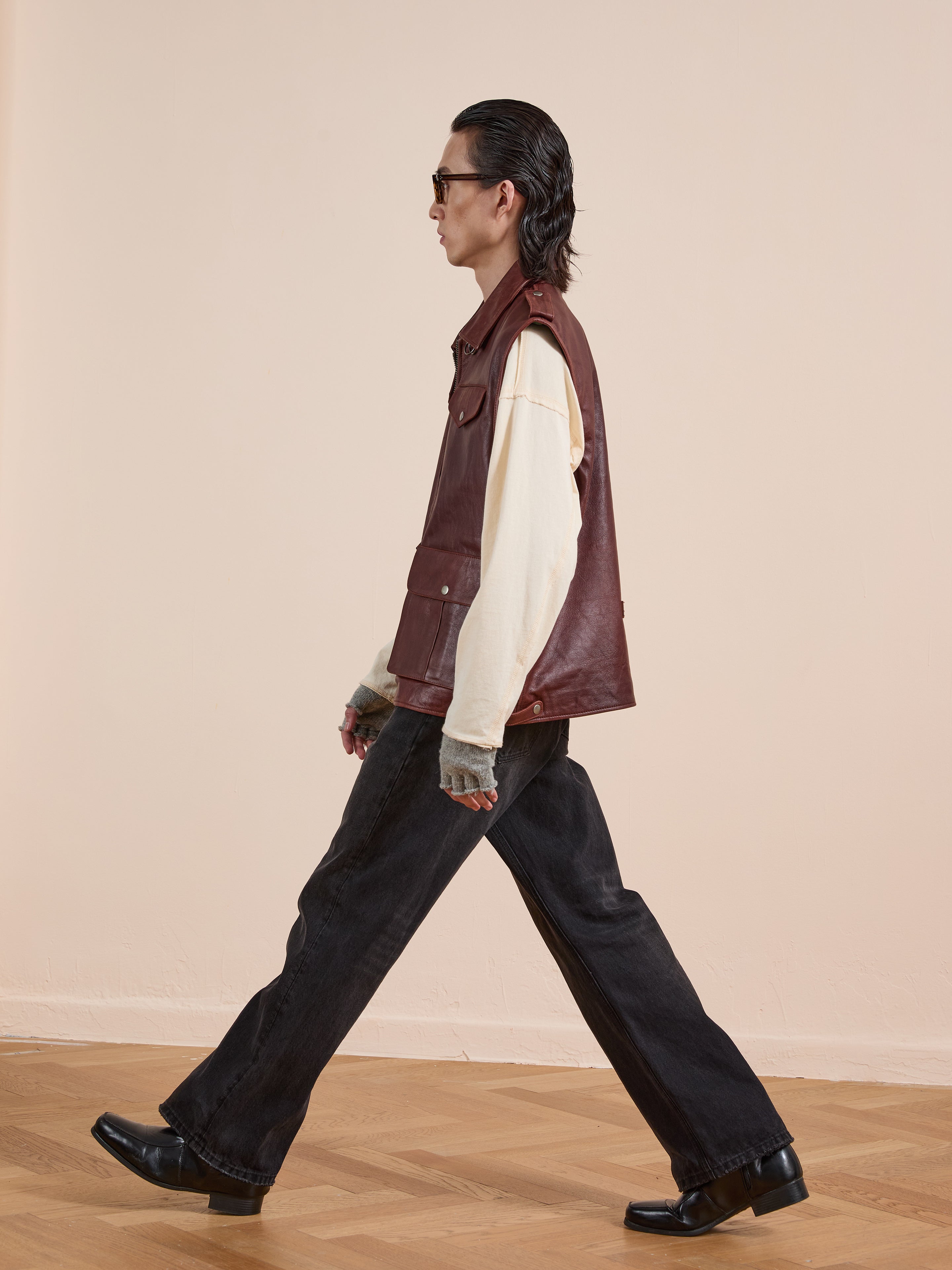 A person with long dark hair, sunglasses, and a Found Leather Pocket Vest over a cream shirt, black wide-leg pants, and black boots walks indoors on a wooden floor.