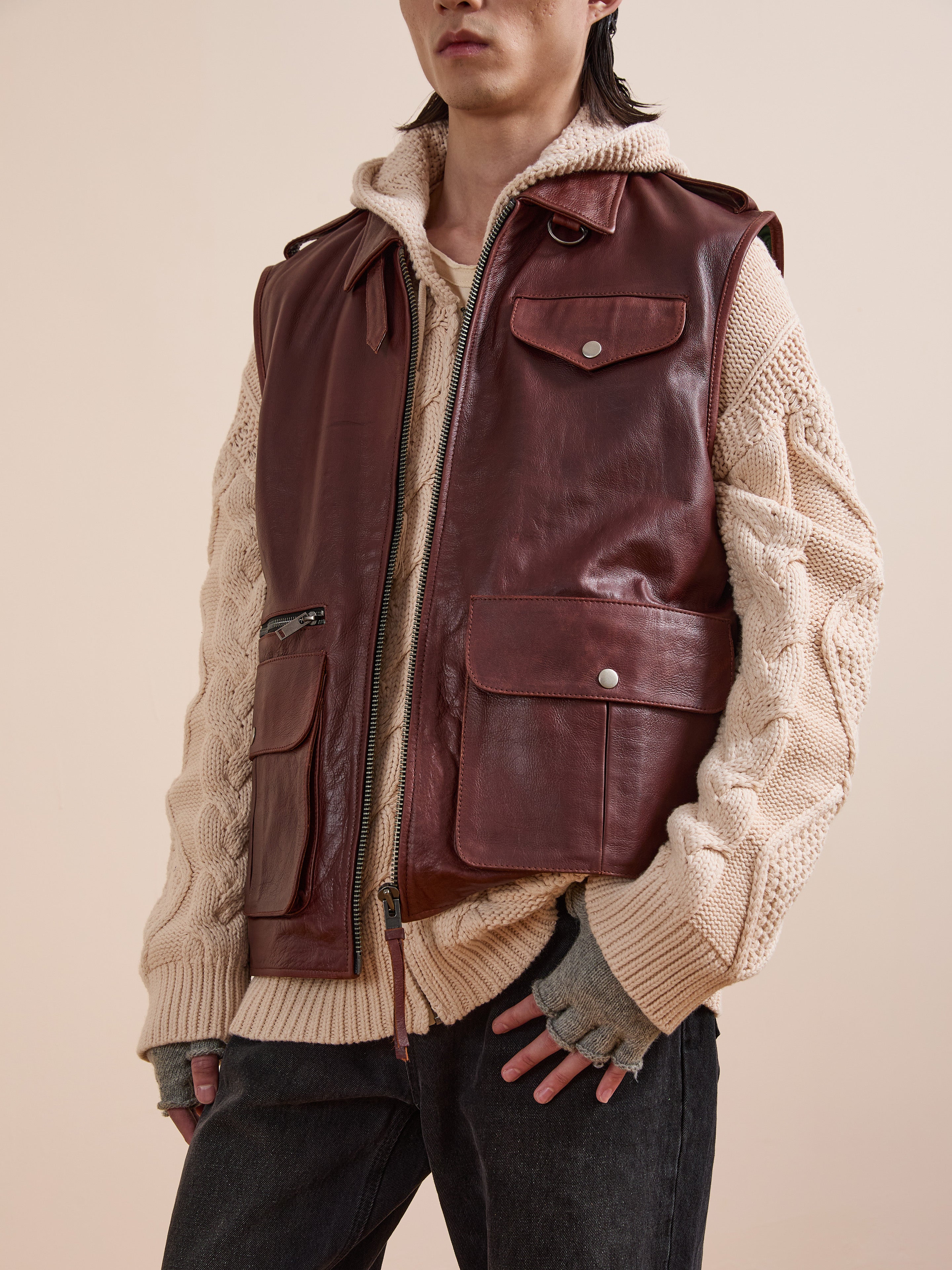 A person wearing Found's Leather Pocket Vest, a chunky cream knit sweater, and dark jeans stands against a beige background.