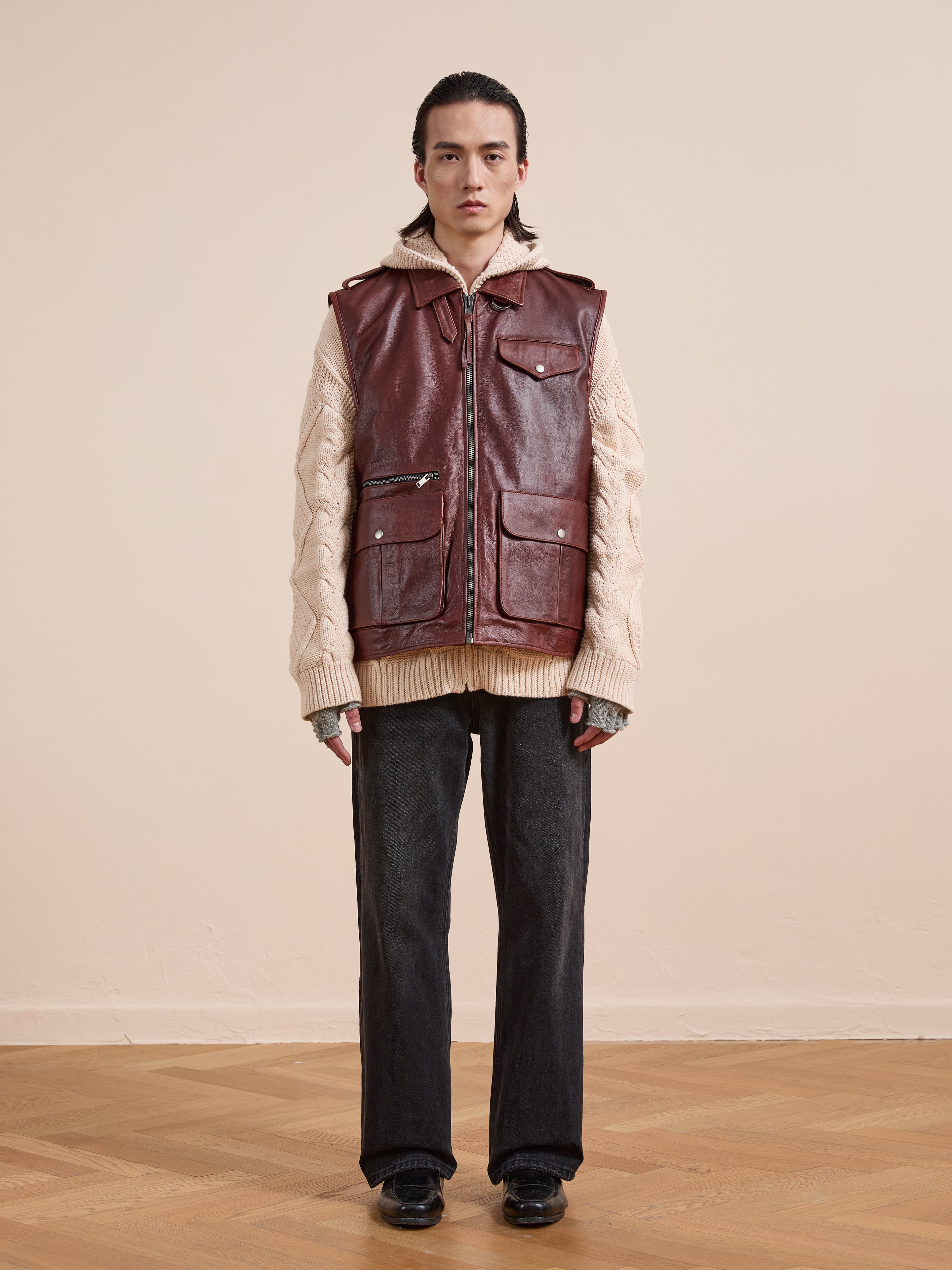 A person stands indoors on a light wood floor against a beige wall, wearing the Found Leather Pocket Vest with utility pockets over a beige cable-knit sweater, paired with dark jeans and black shoes.