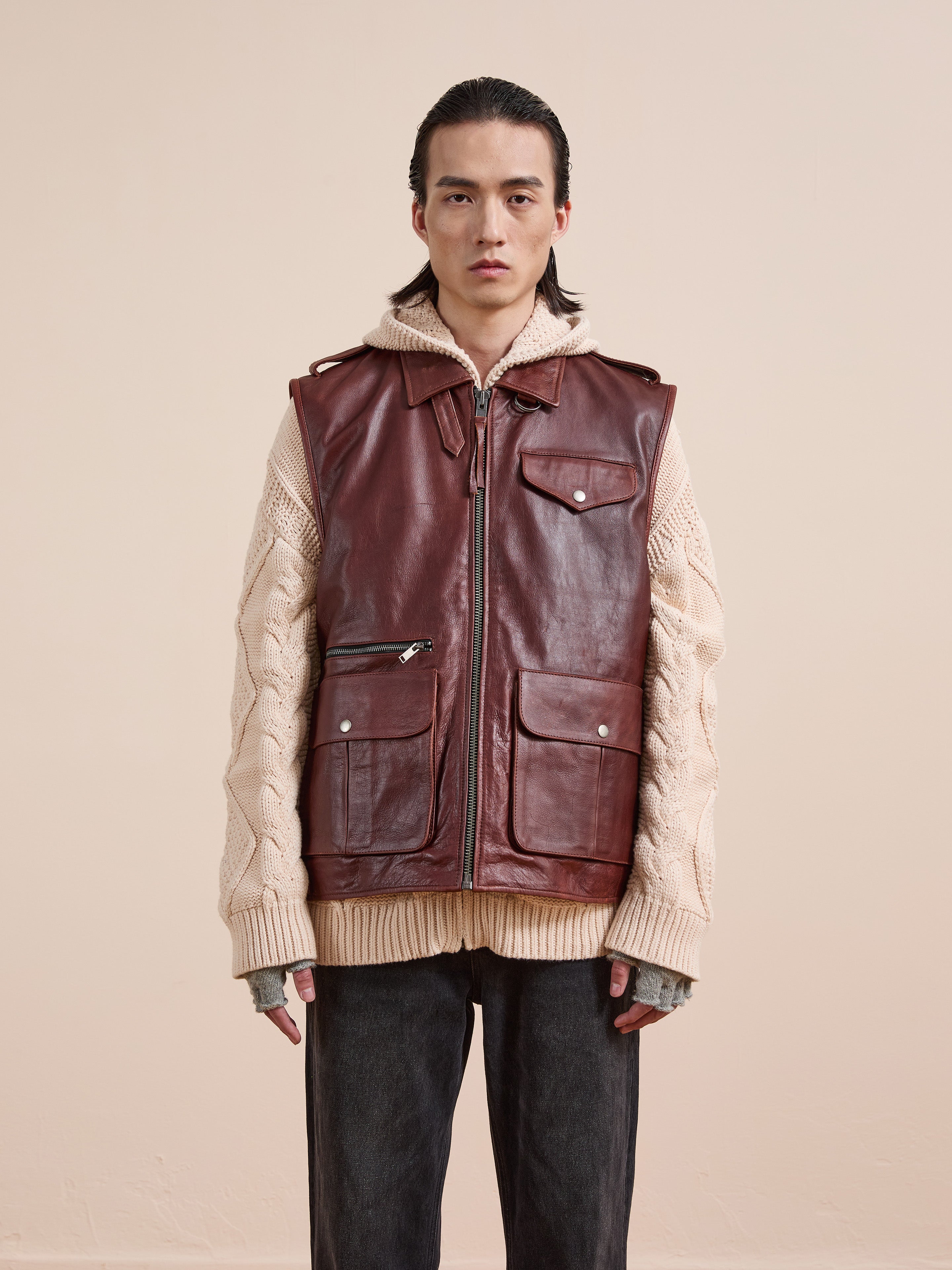A person stands against a plain background wearing a cream cable-knit sweater, dark pants, and the Found Leather Pocket Vest.