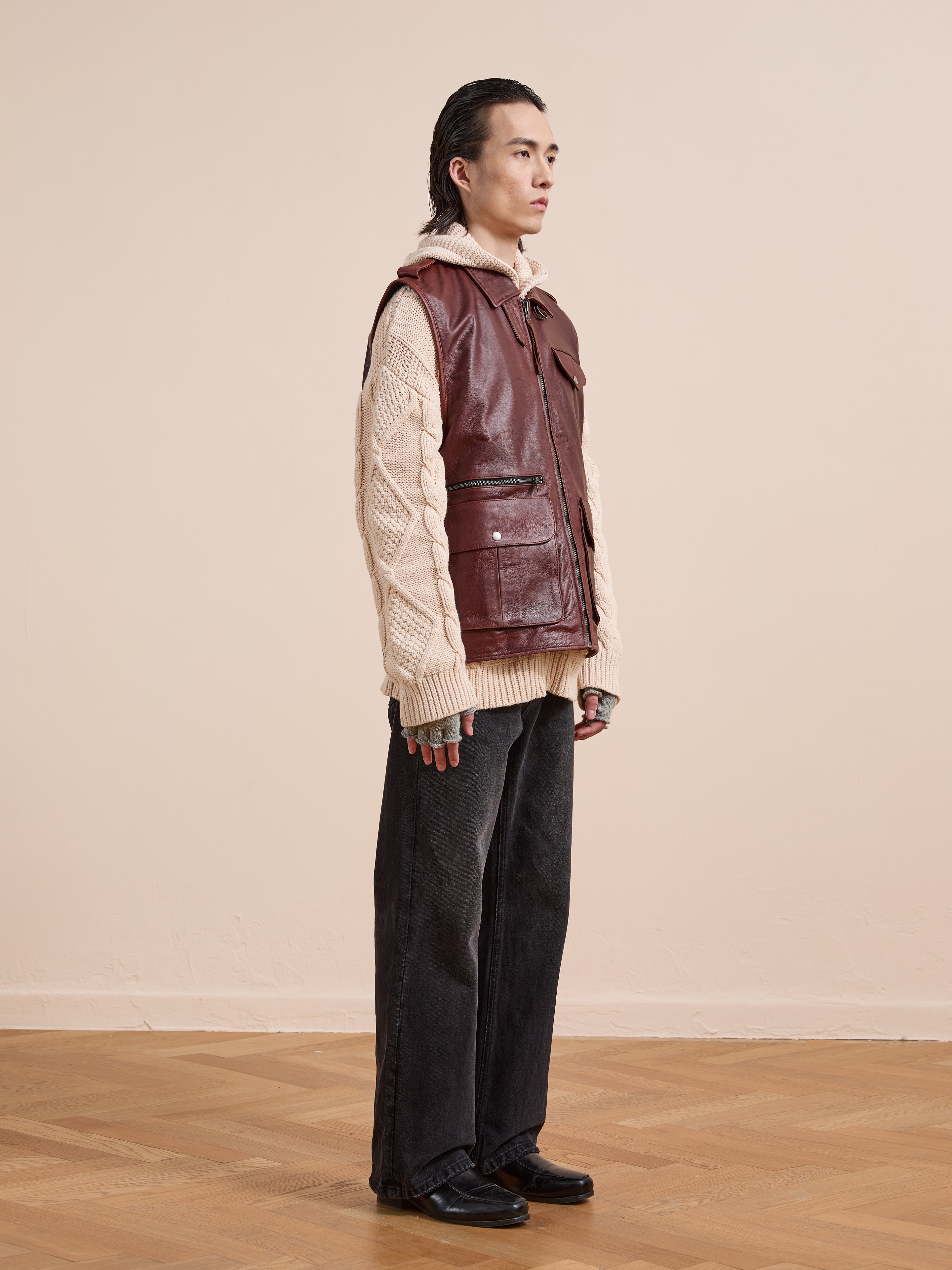 A person stands indoors against a beige background wearing the Found Leather Pocket Vest in maroon over a cream cable-knit sweater, paired with loose black jeans and black shoes on a wooden floor.