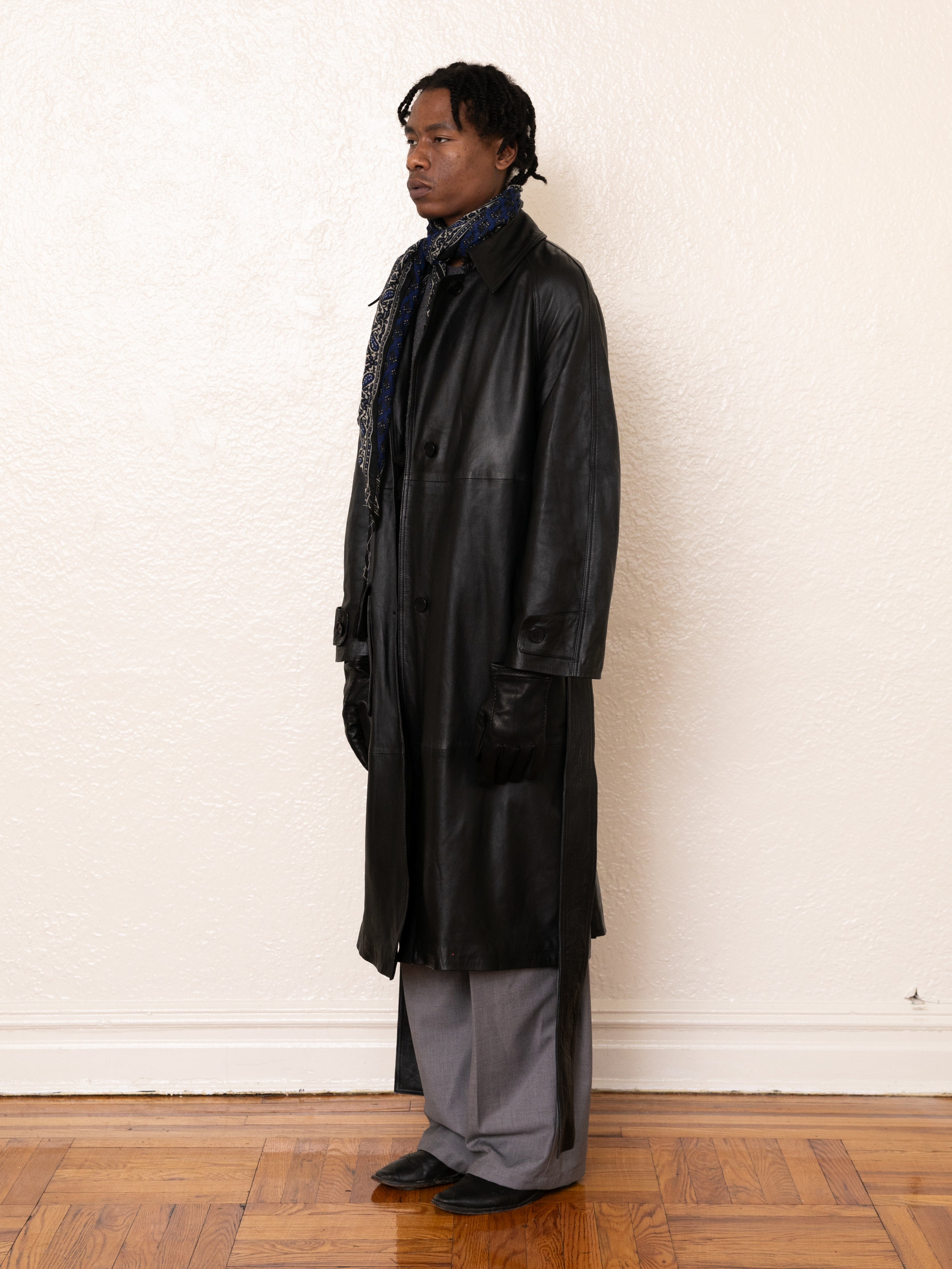 A person in a FOUND ethically crafted Leather Trench Coat, gray pants, scarf, and gloves stands on a wooden floor against a plain wall.